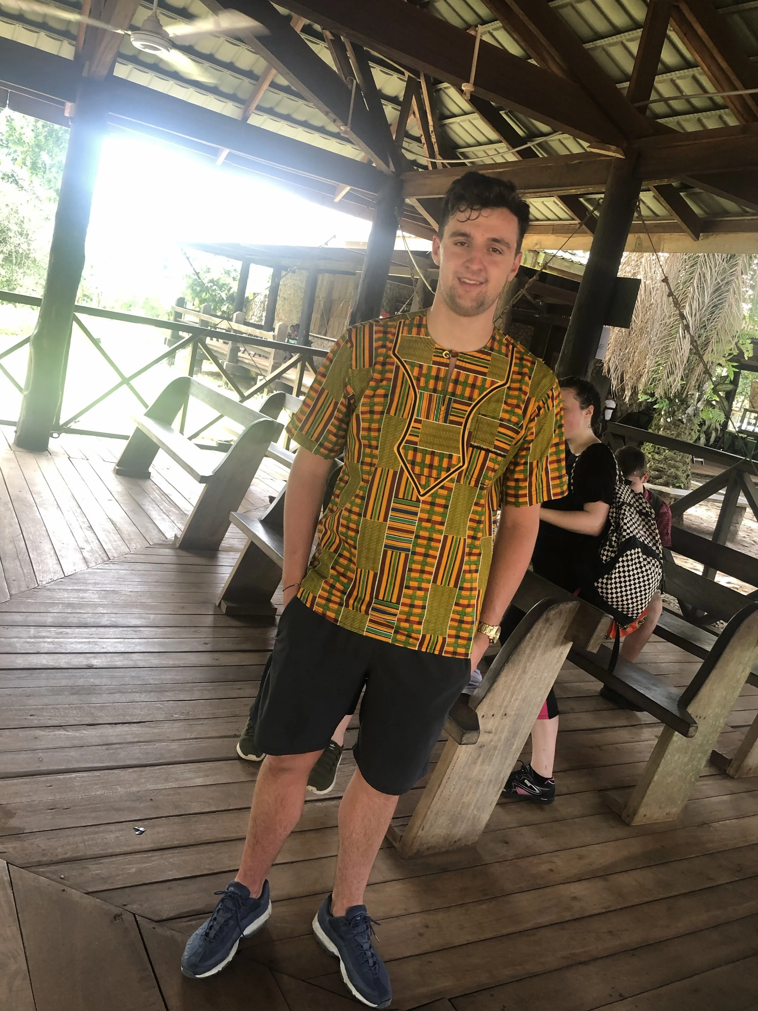 A young man in a colorful African pattern shirt and black shorts posing indoors on a wooden bench in a rustic setting with a high ceiling and wooden beams, with other people visible in the background.