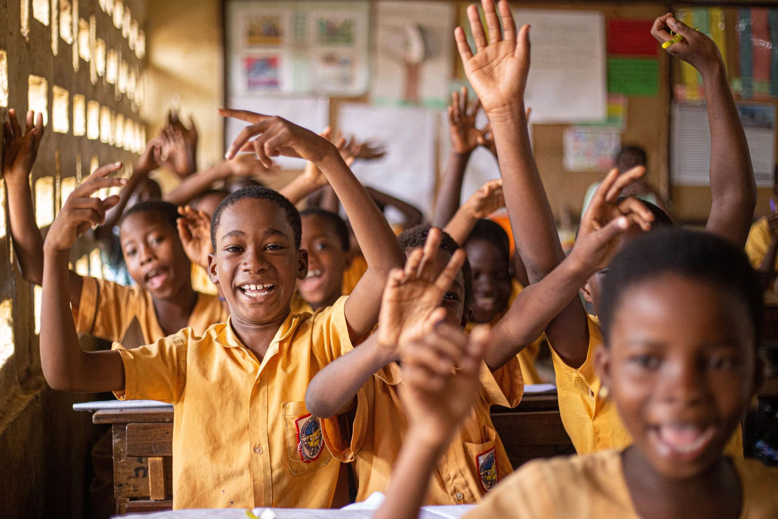 Children in yellow school uniforms are raising their hands inside a classroom, smiling and engaging in a question or activity.
