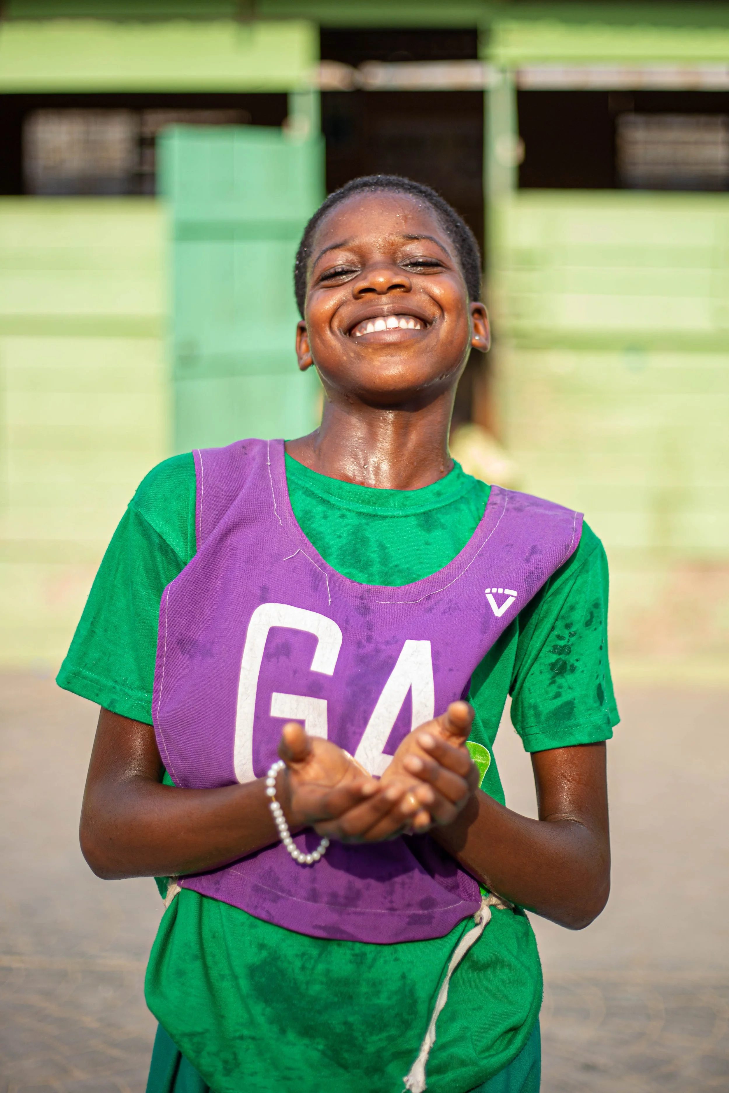 A smiling young girl with short hair, wearing a green shirt and purple athletic bib with the number 64, standing outdoors in front of a background of green and brown structures, clapping her hands.