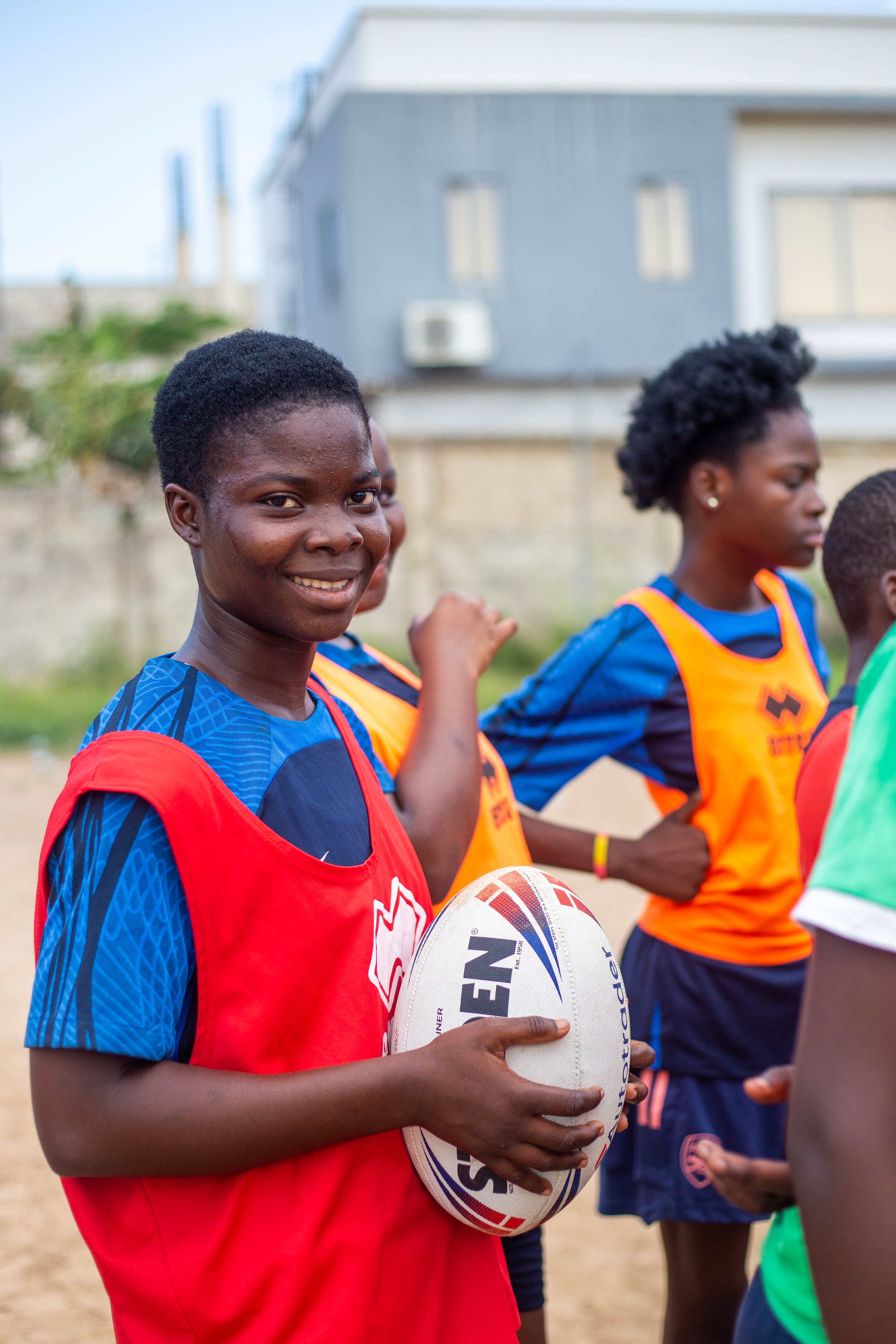 A young girl with short hair wearing a red and blue sports jersey holding a rugby ball during a match or practice session on a field, with other players in similar jerseys in the background.