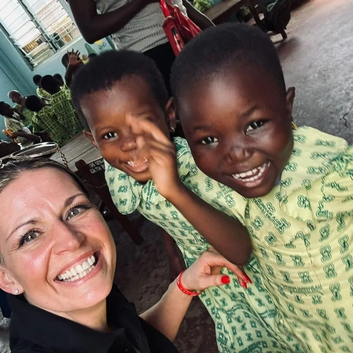 Smiling woman taking a selfie with two young boys in school uniforms, all looking happy. Background shows other children and classroom setting.