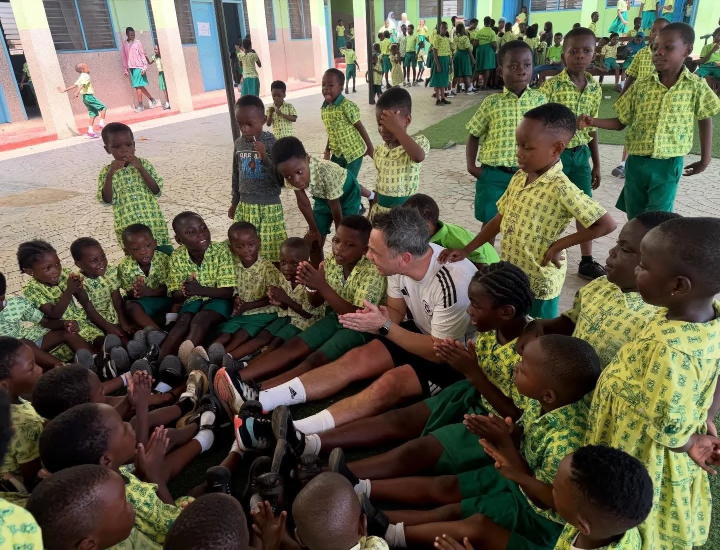 A group of young school children in green and yellow uniforms gathered in a classroom or courtyard, sitting and standing around a man who is talking and clapping.