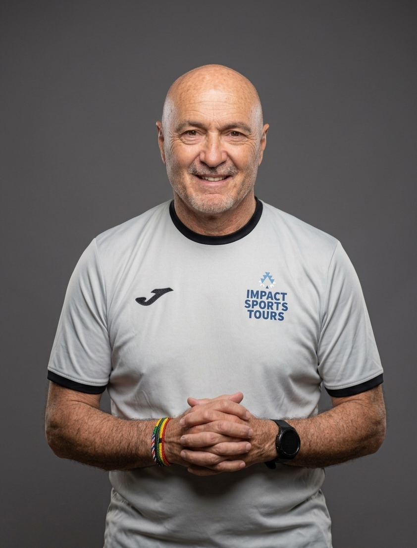A smiling older man with a shaved head and gray beard, wearing a white sports shirt with a logo that says 'Impact Sports Tours,' standing against a gray background.