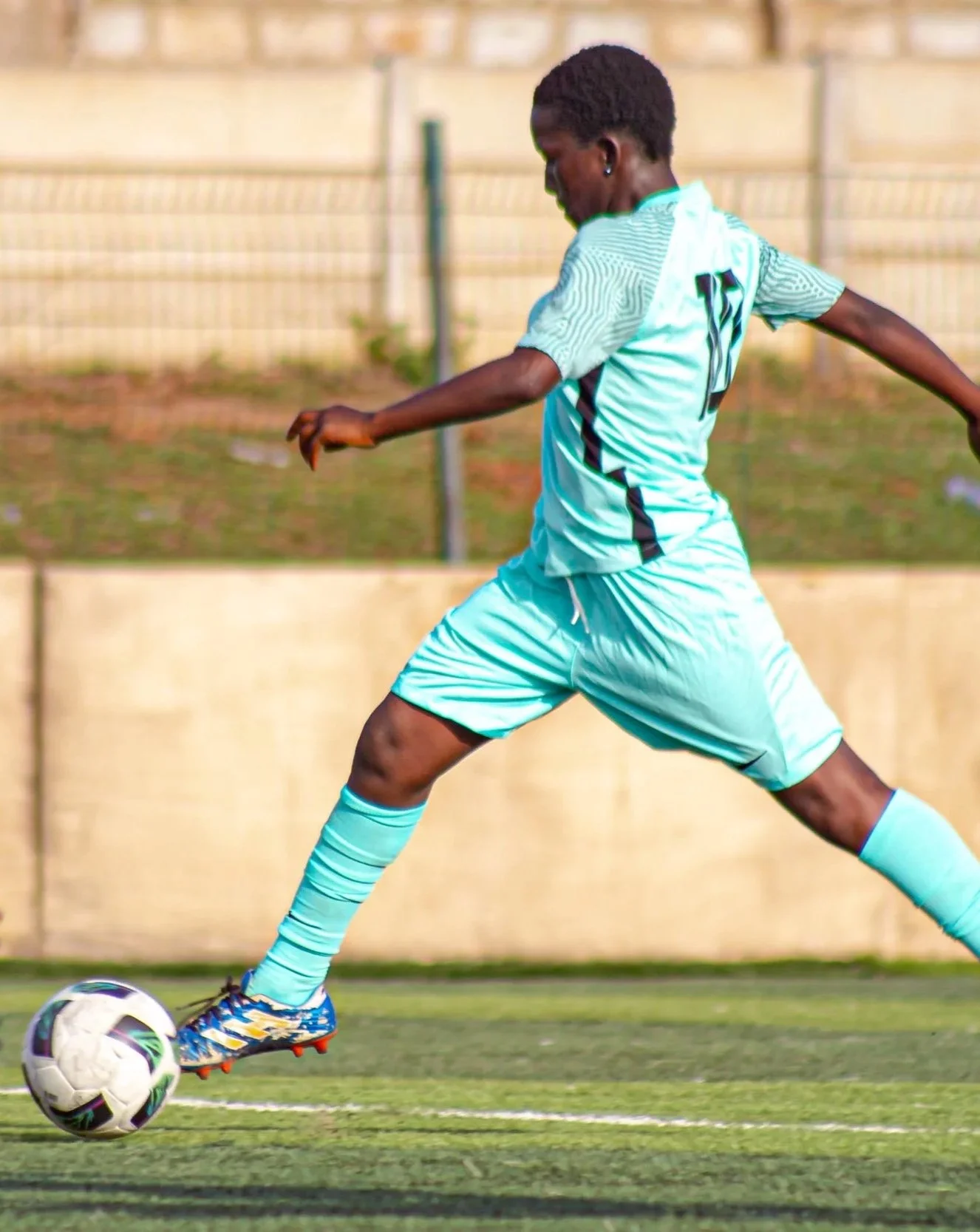 A male soccer player is in motion, kicking a soccer ball on a field, wearing a light blue uniform with the number 12 on the back.