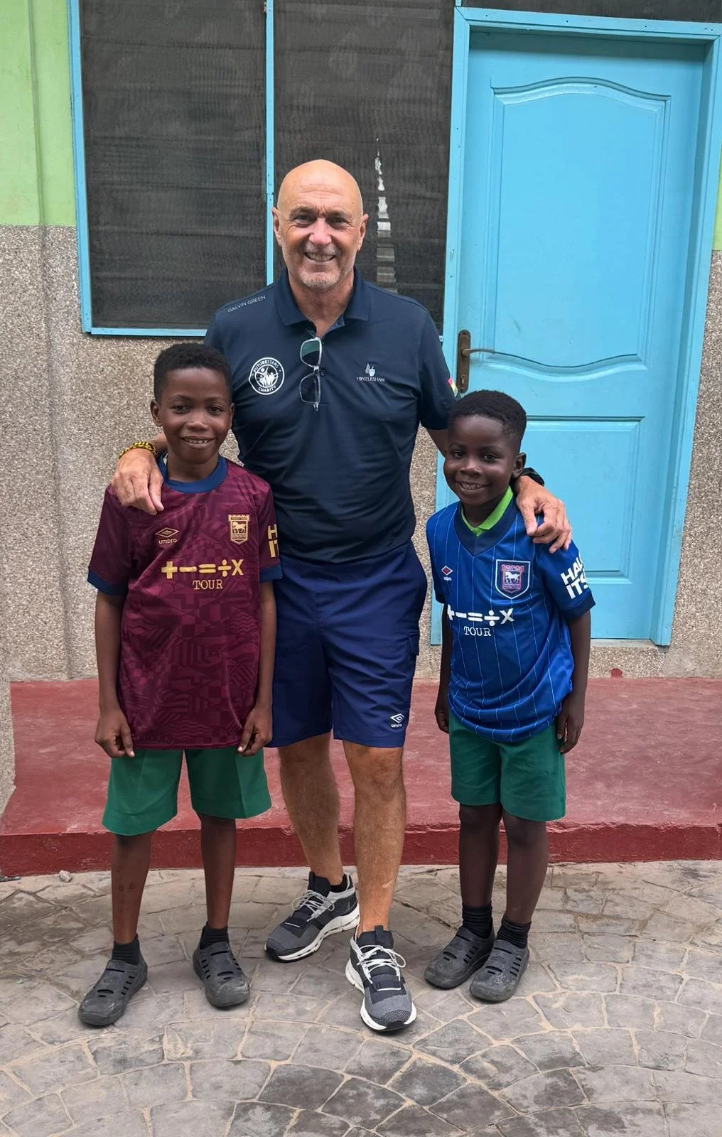 A man wearing a navy blue polo shirt and shorts, poses with two young boys. The boys are smiling, one wearing a maroon soccer jersey and green shorts, the other in a blue soccer jersey and green shorts. They are standing outside in front of a light blue door with a textured concrete wall behind them.