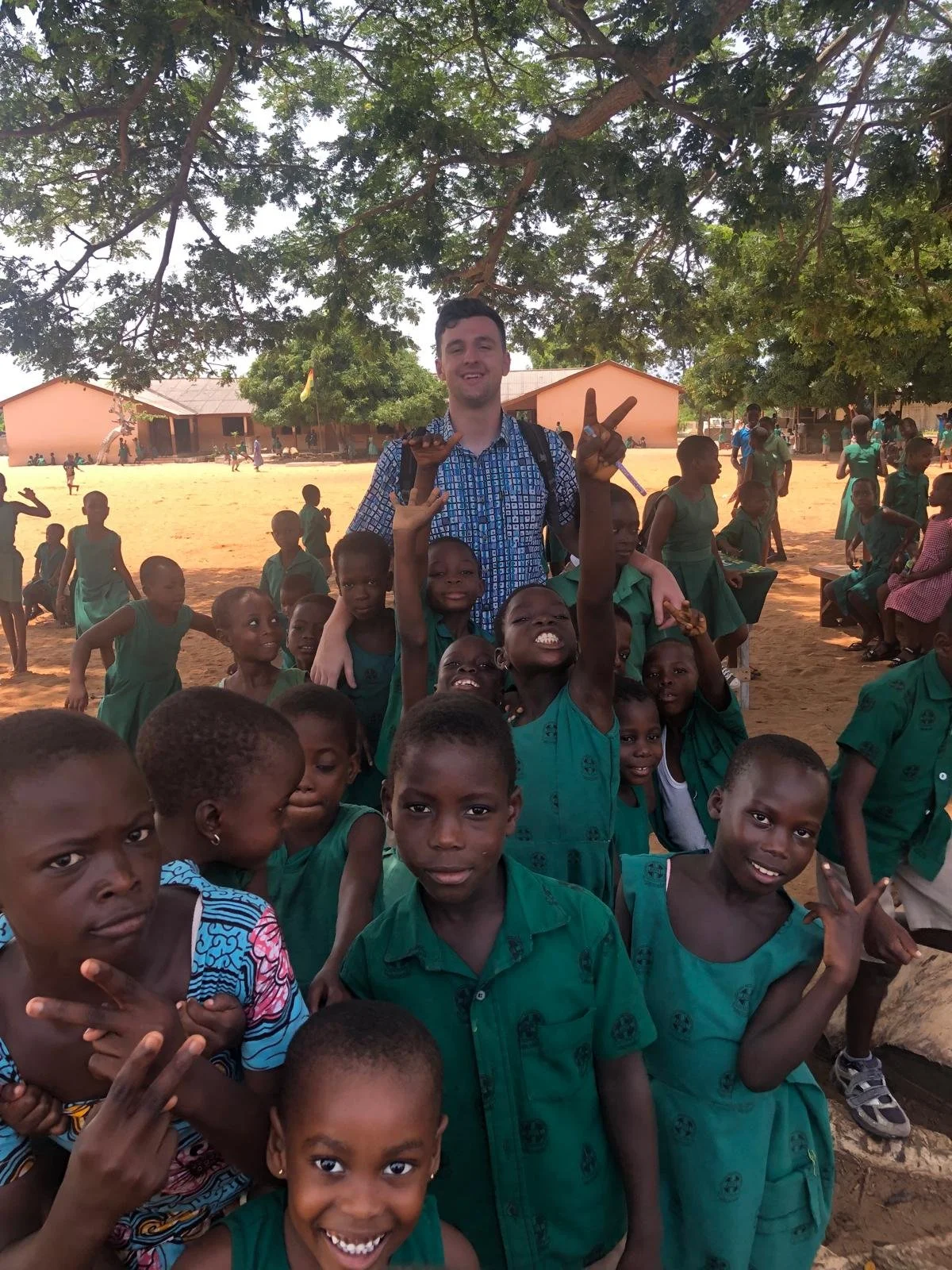 A group of children in school uniforms outside on a dirt field, with a young man among them making peace signs, under large trees with buildings in the background.