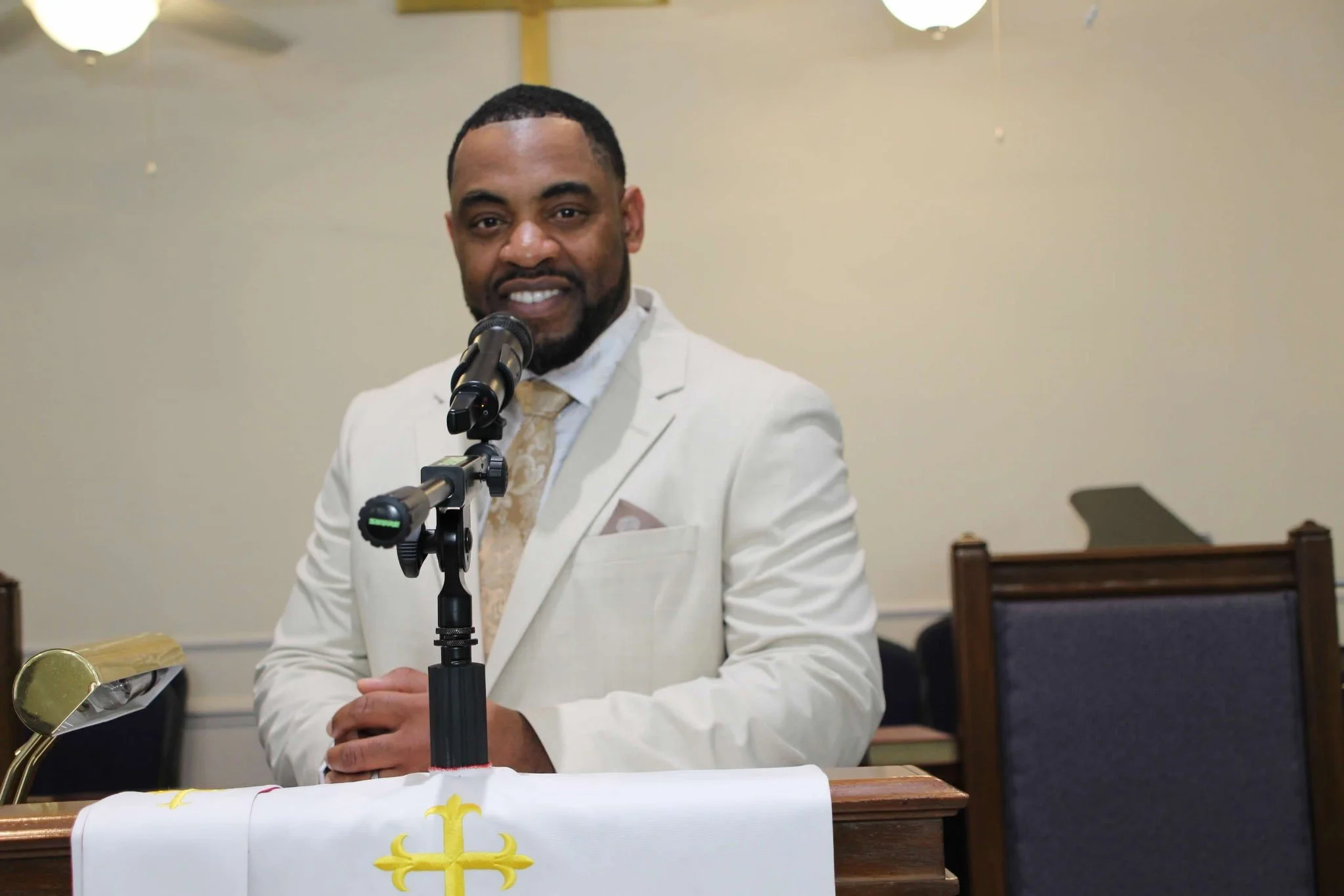 A man dressed in a white suit is smiling, standing behind a microphone, in a room with beige walls and ceiling lights.