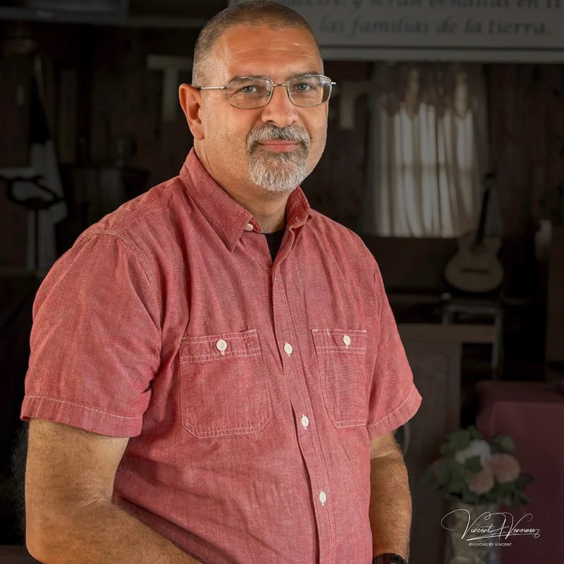 A middle-aged man with glasses, a short beard, and a buzz cut, wearing a red short-sleeve button-up shirt, standing indoors in front of a background with musical instruments and a flower arrangement.