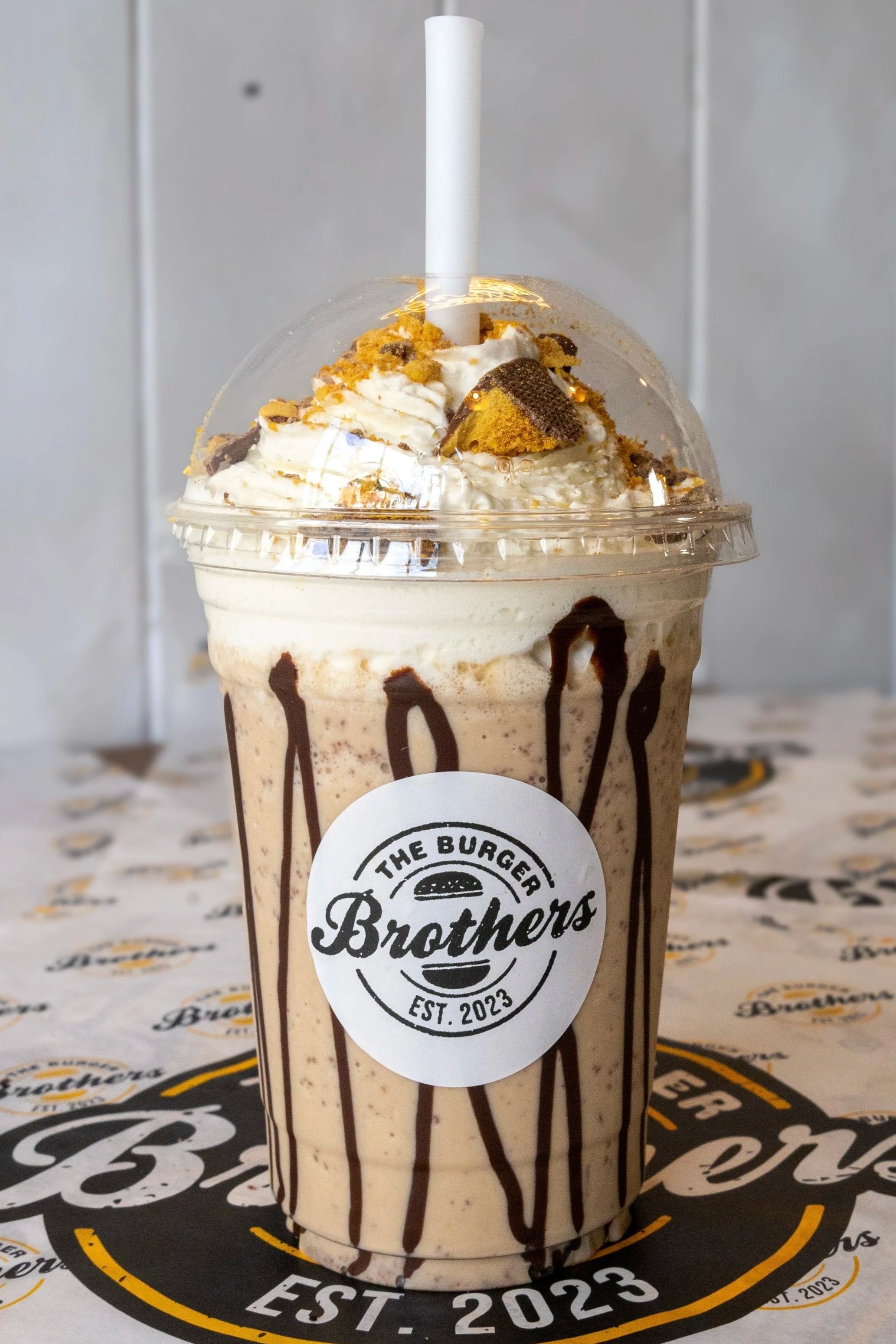 A blended chocolate and vanilla milkshake topped with whipped cream, cookie crumbles, and chocolate drizzle in a branded cup with a straw, placed on branded paper and background.