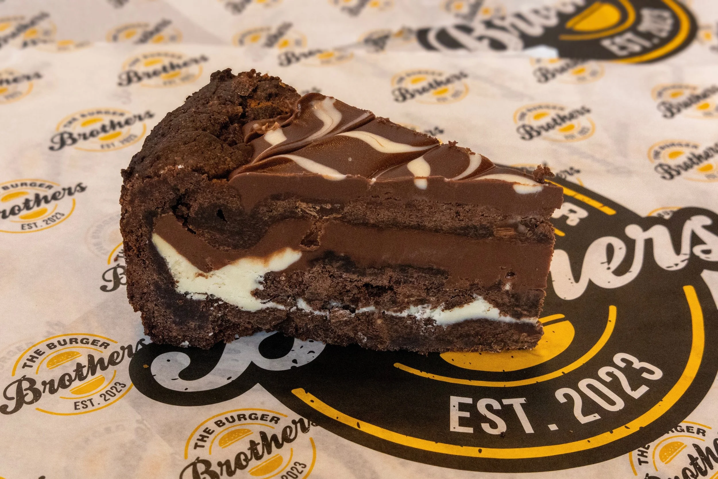 A slice of chocolate brownie with a marbled chocolate and white topping on branded parchment paper from The Brothers burger establishment.