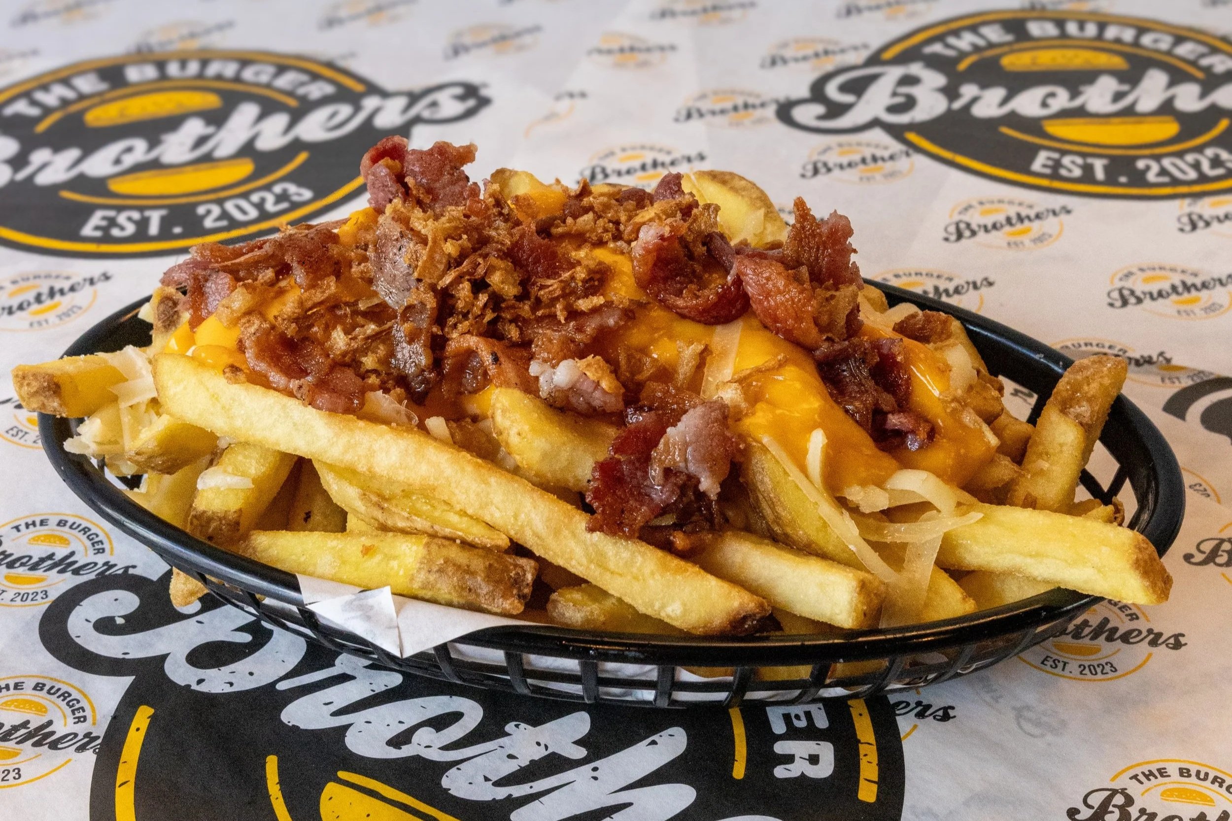 Basket of French fries topped with cheese, bacon bits, and shredded cheese, placed on a tray with printed paper.
