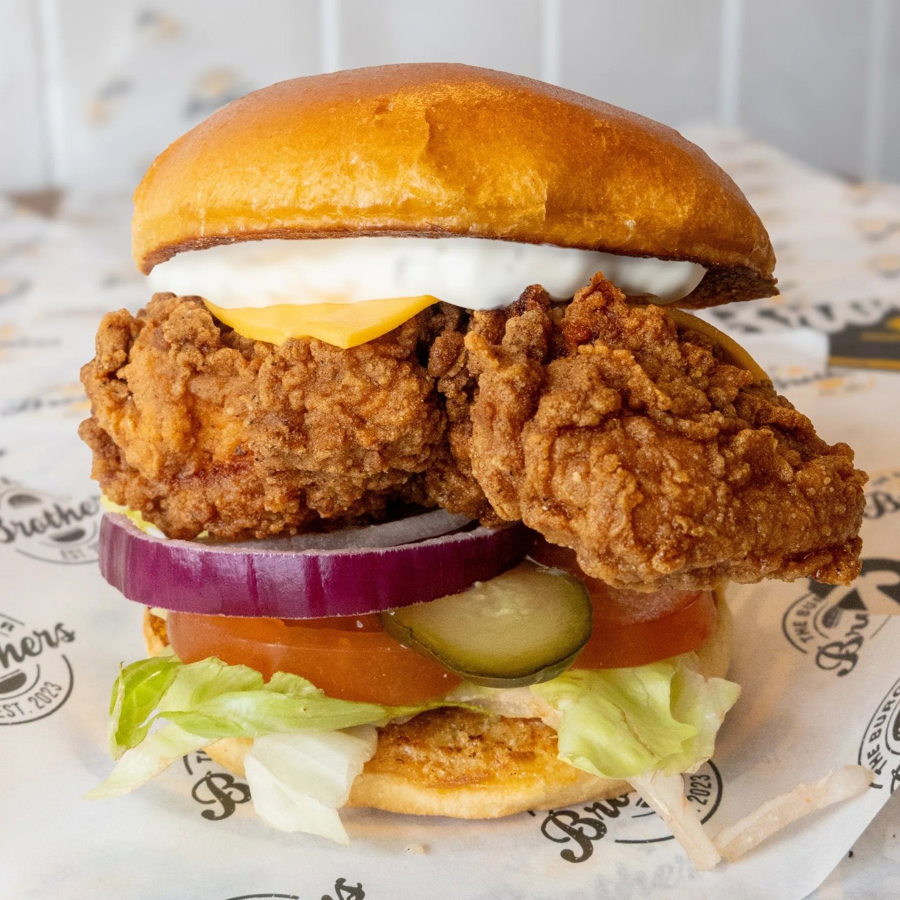 Close-up of a fried chicken sandwich with pickles, tomato, lettuce, cheese, mayonnaise, and a toasted bun.