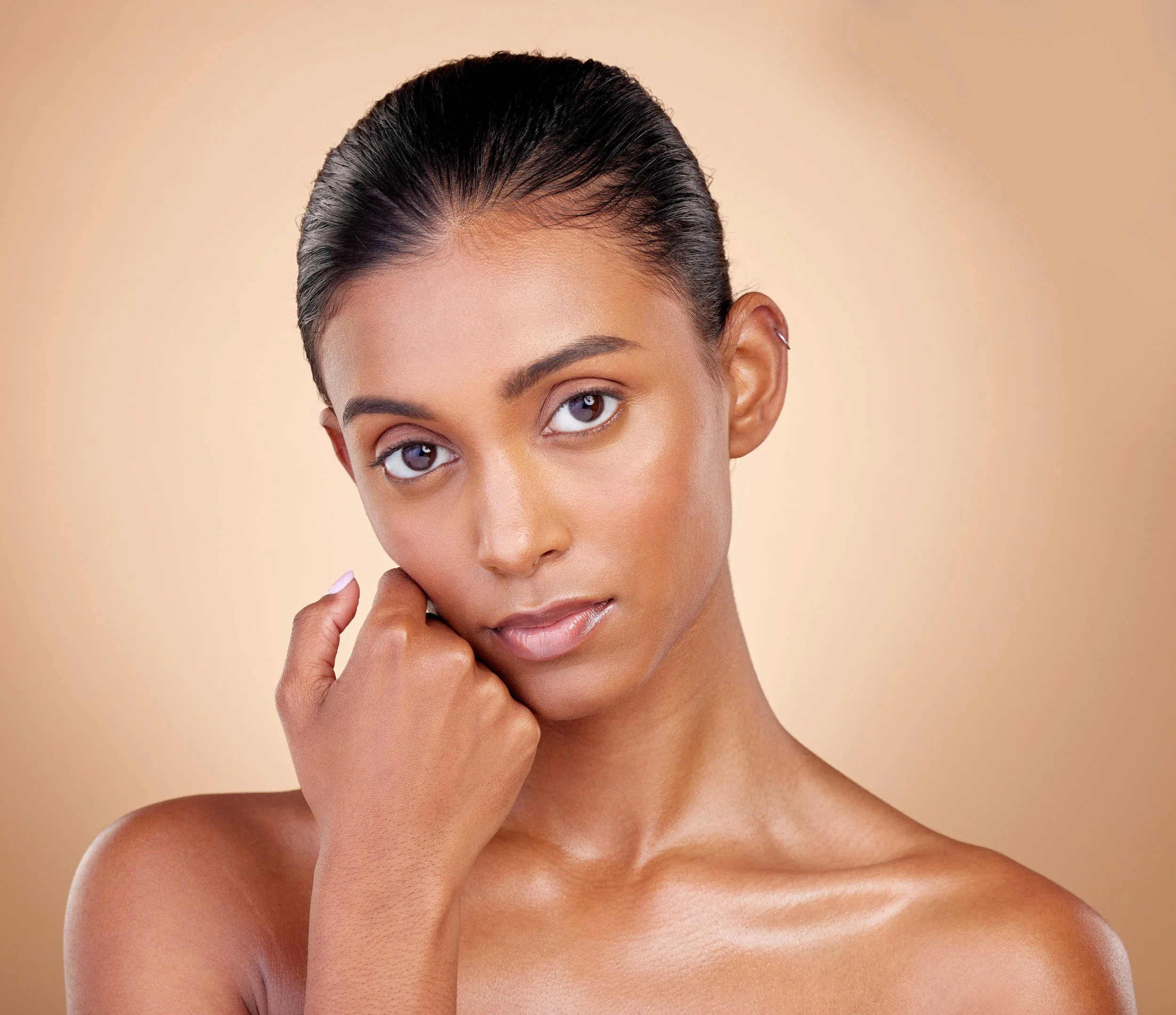 Close-up of a woman with dark, wet hair slicked back, touching her chin with her hand, against a neutral beige background.