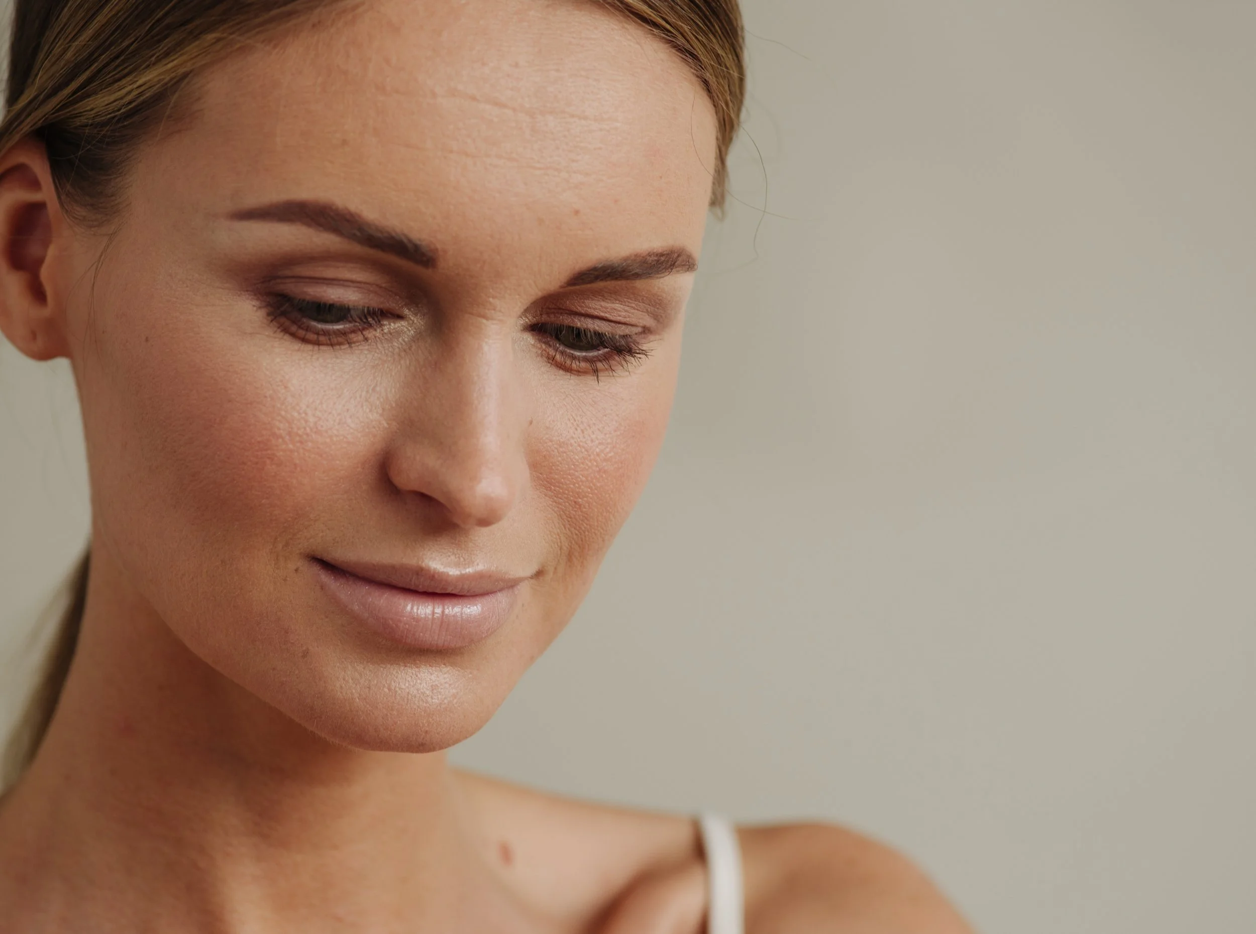 Close-up of a woman with fair skin, reddish-brown hair, and makeup, looking slightly downward. She is wearing a white strap on her shoulder.