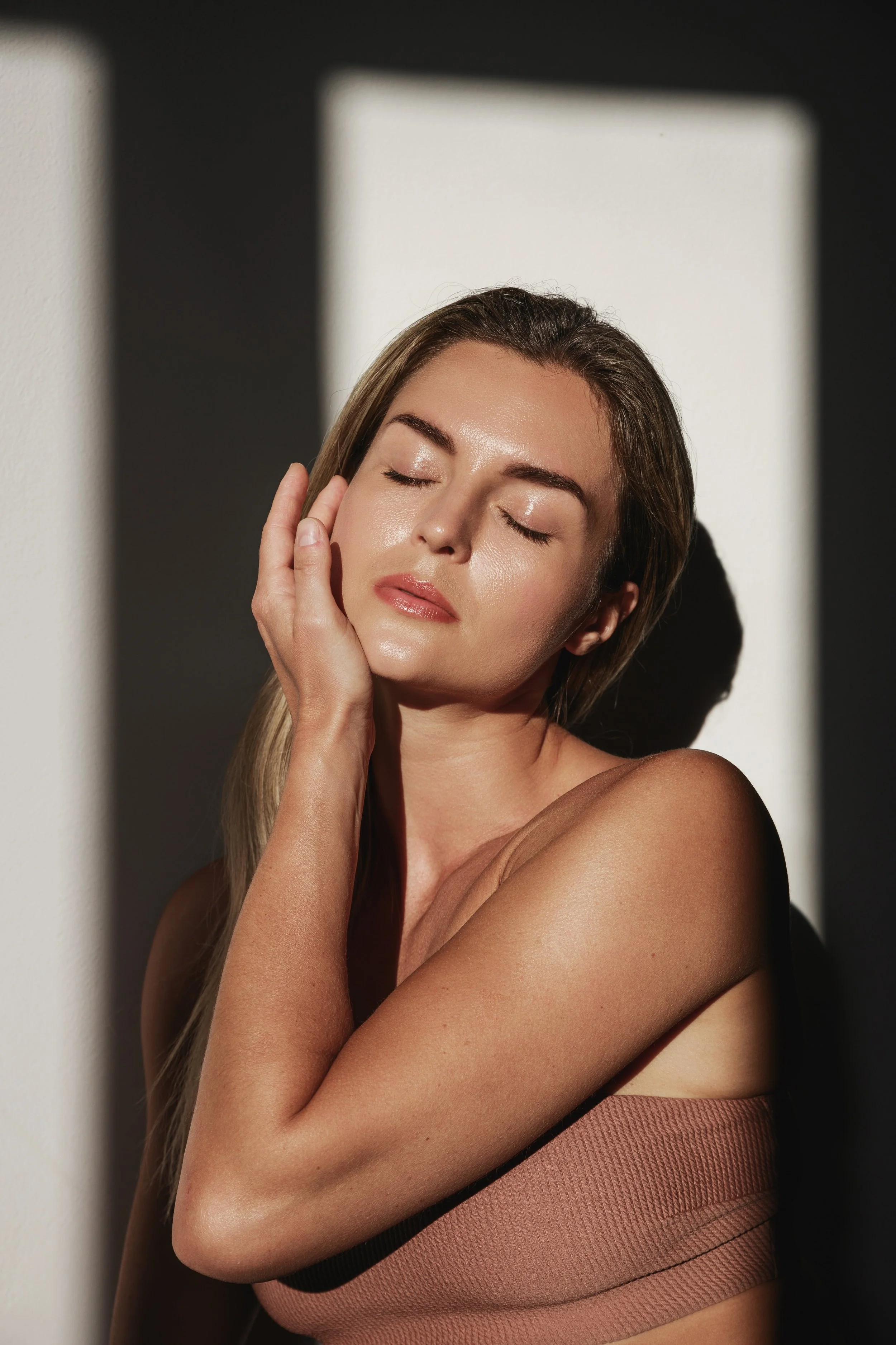 A woman with closed eyes and a relaxed expression touches her cheek with her hand. She has long, straight hair and smooth skin. She is wearing a sleeveless, textured, pinkish-beige top. The background features soft shadows and light patterns on a wall.