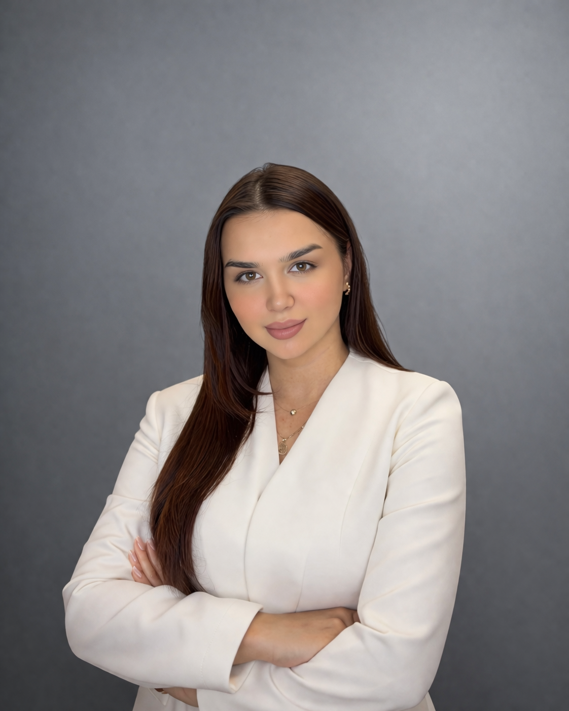 A portrait of a woman with long brown hair, wearing a white blazer, standing with arms crossed against a gray background.