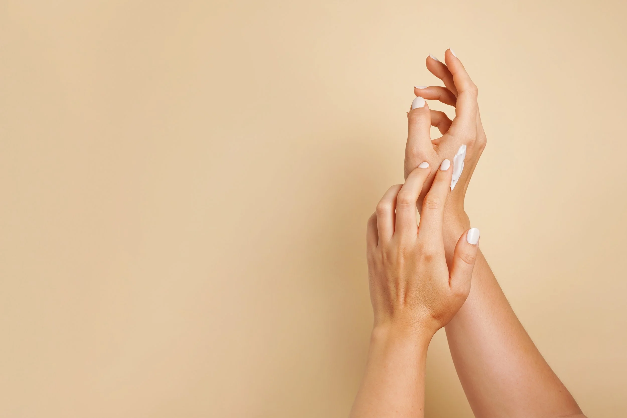 Hands with white nail polish applying lotion or cream to the skin against a beige background.