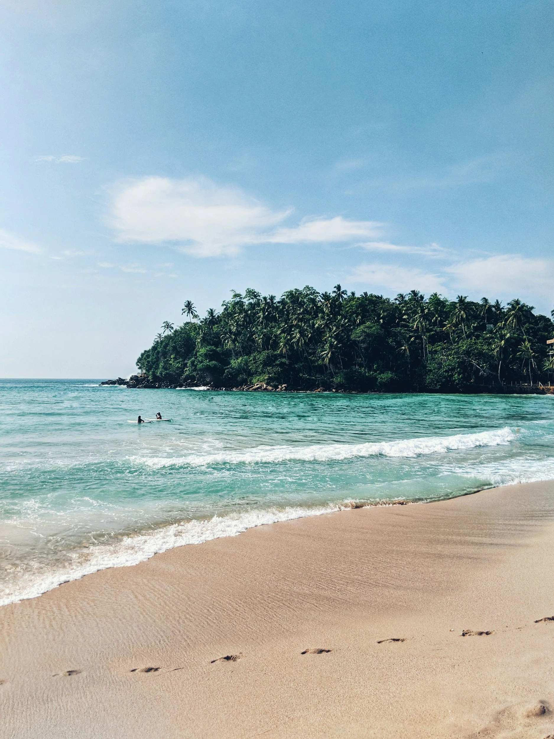 A sandy beach with footprints, greenish waves, a rocky shoreline, a lush green hill with palm trees, and a clear blue sky with some clouds.