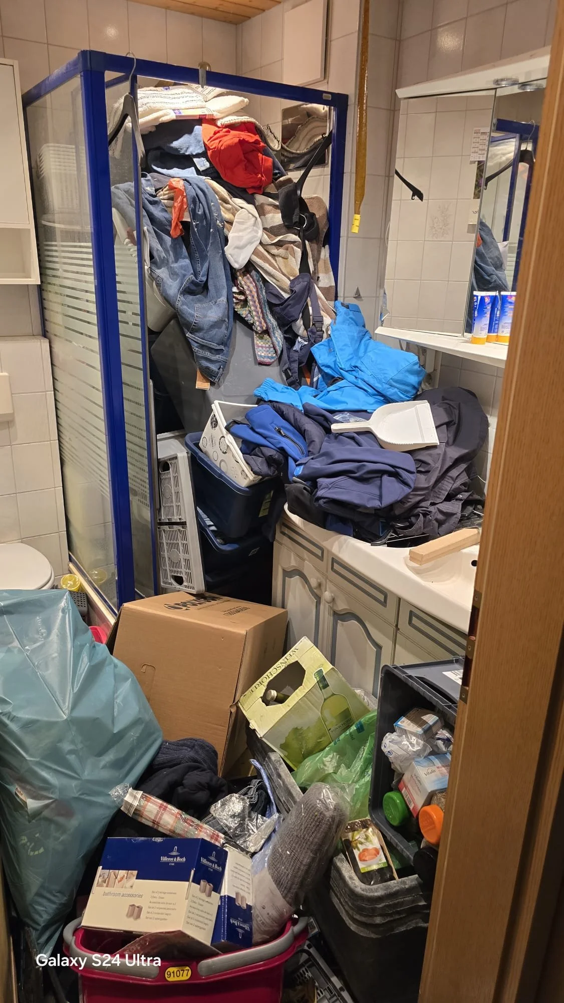 Ein unordentlicher Raum mit Kleidung, Papiertüten, Kartons, Reinigungsmitteln, einem Spiegel, einem waschbecken und anderen Haushaltsartikeln.
