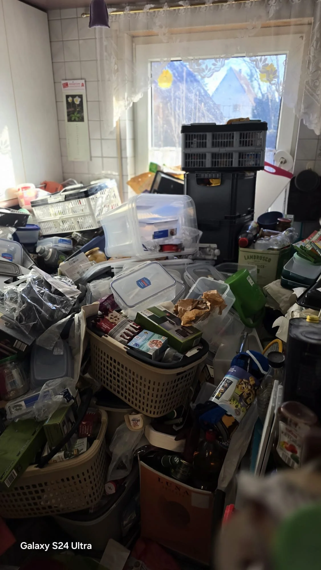 Ein unordentlicher Raum voller Müll, Plastikbehälter, Verpackungen und Haushaltsgegenstände vor einem Fenster mit geformten Vorhängen.