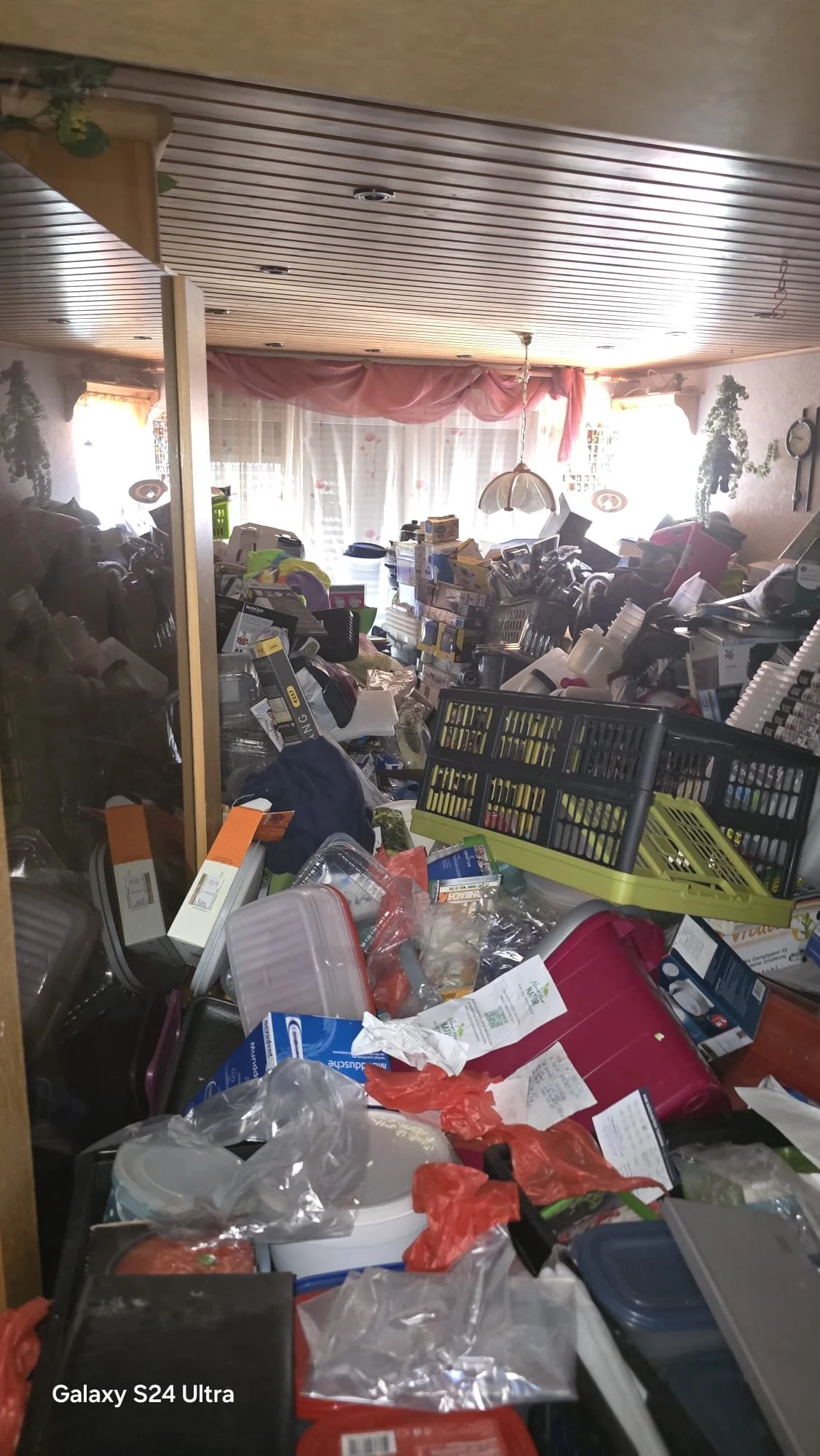 Ein unordentlicher Raum mit zerstreuten Gegenständen, Kartons, Verpackungen und Haushaltsartikeln. Im Hintergrund sind Fenster mit Vorhängen, die viel Licht ins Zimmer lassen.