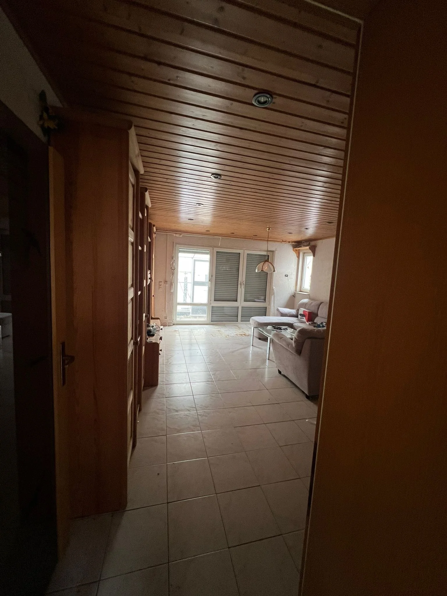 Ein Wohnzimmer mit Holzboden und Holzdecke, große Fenster mit Rollläden, Sofa, Couchtisch und Regal.