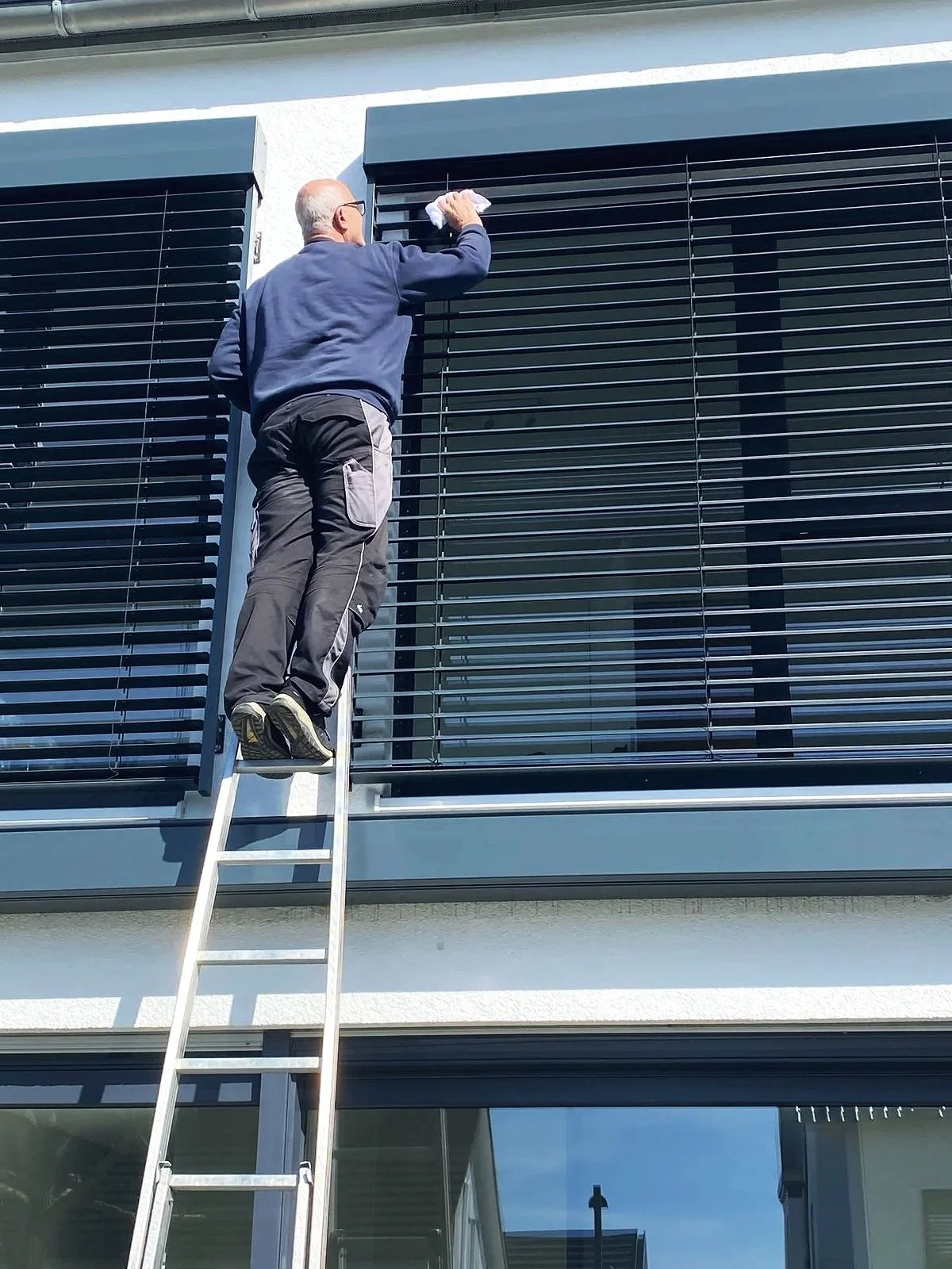 Ein Mann steht auf einer Leiter vor einem großen Fenster mit Rollläden und wischt sie ab.