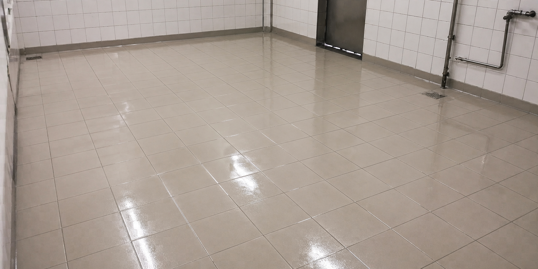 Boden im Badezimmer mit weißen Fliesen, Wasserflecken und Abfluss. Weiße Wandfliesen, Rohrleitungen an der Wand und eine Metalltür im Hintergrund.