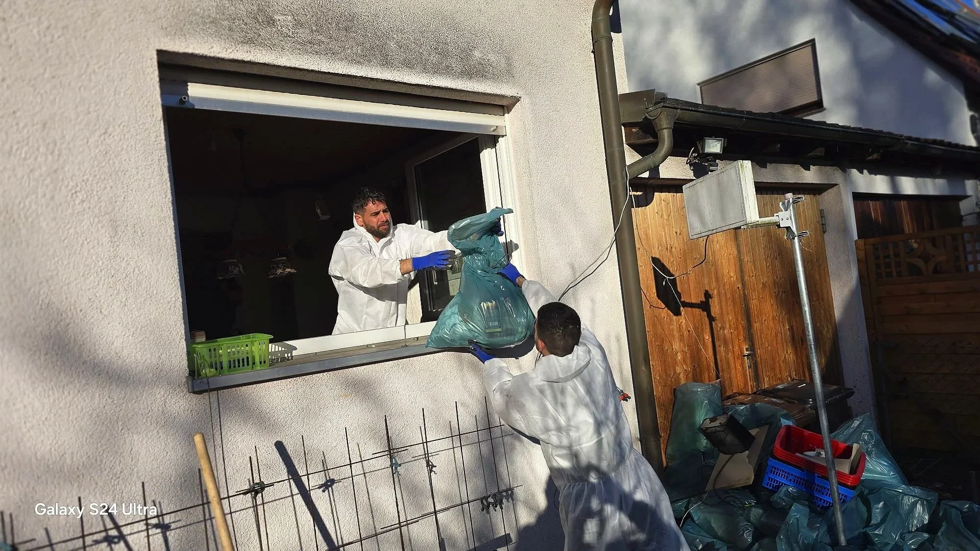 Zwei Männer im weißen Schutzanzug, die Pakete durch ein Fenster einer weißen Hauswand übergeben, während sie draußen stehen. Es gibt eine Holzterrasse, eine Bauleine, Werkzeuge und blaue Plastikbeutel im Vordergrund.