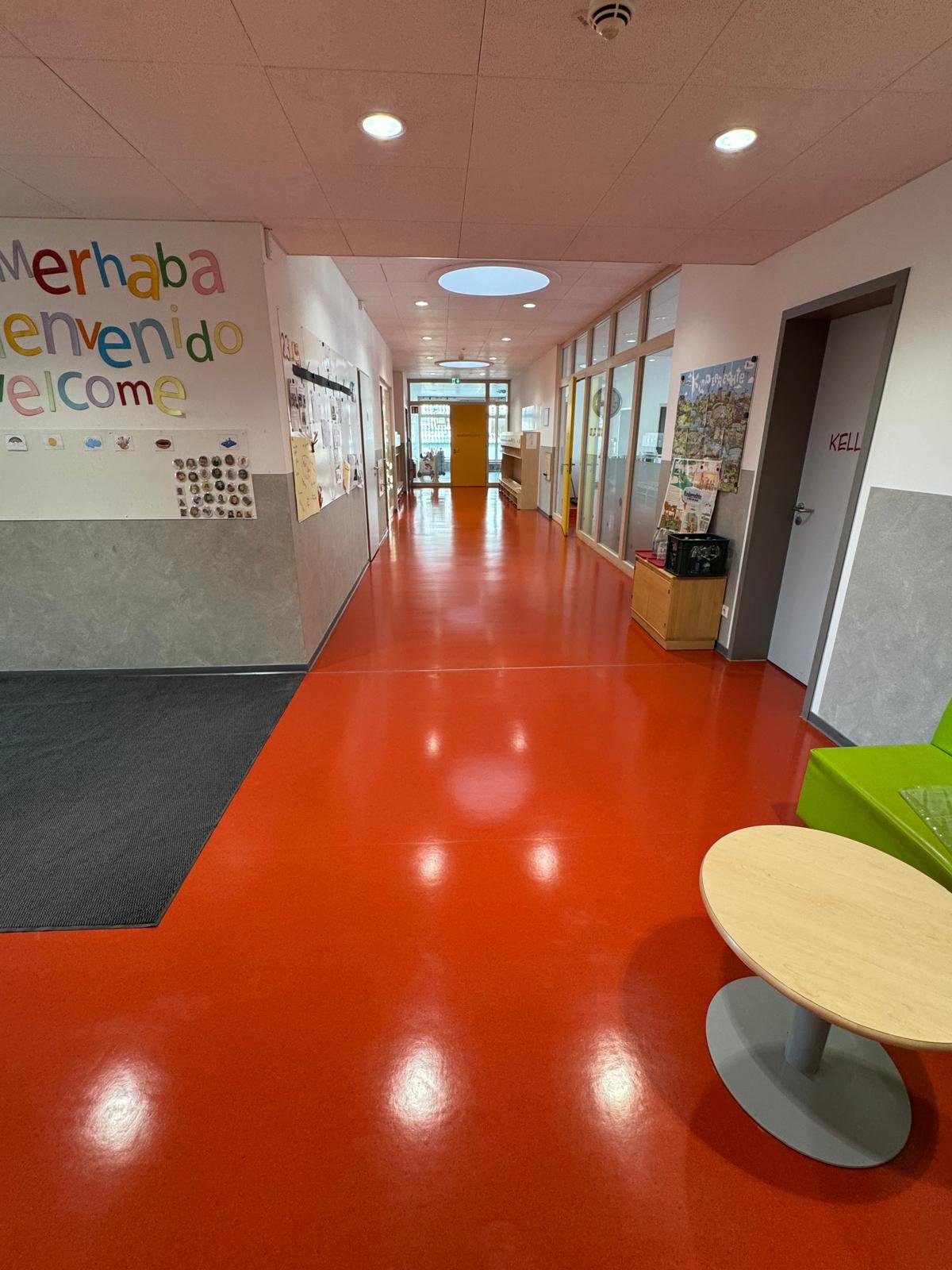 Heller Flur in einer Kindertagesstätte oder Schule mit orangefarbenem Boden, weißen Wänden, einigen Dekorationen und einem Wartebereich mit kleinen Sitzgelegenheiten.