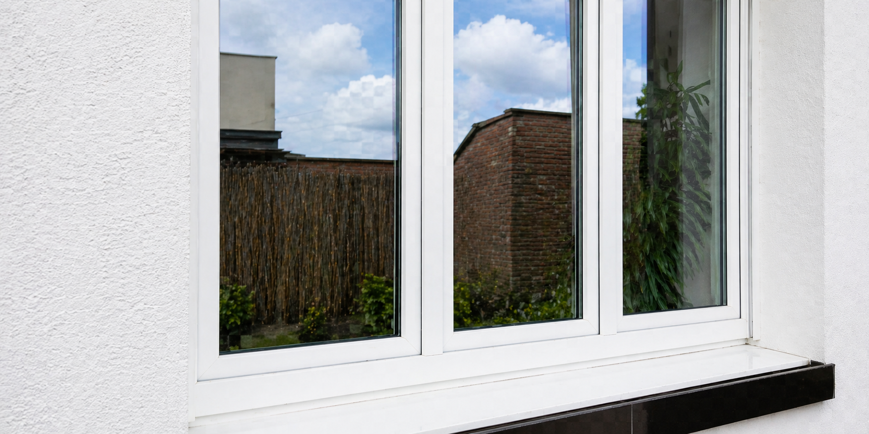Ein Fenster mit weißem Rahmen an einer weißen Wand, Blick nach draußen auf einen Garten mit Pflanzen, Zaun und Gebäuden im Hintergrund.
