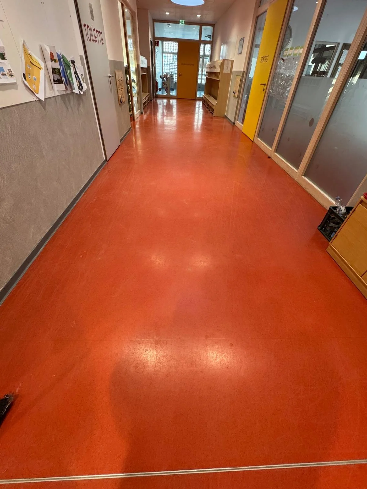Ein leerer Flur in einem Kindergarten oder Vorschule mit rotem Boden, hellem Holzregalen und bunten Türen, die mit Glasfenstern versehen sind.
