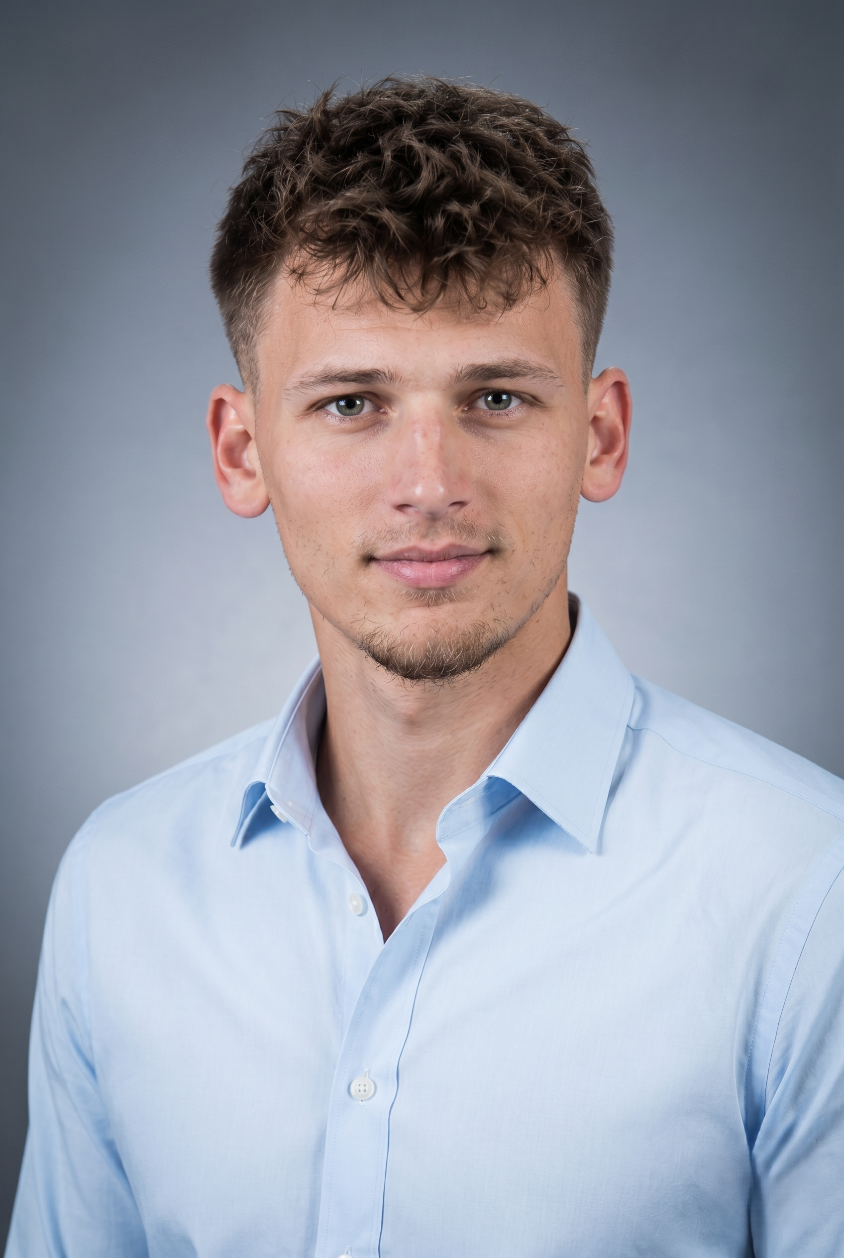 Porträt eines jungen Mannes mit kurzen, lockigen braunen Haaren und blauen Augen, trägt ein hellblaues Hemd vor einem grauen Hintergrund.