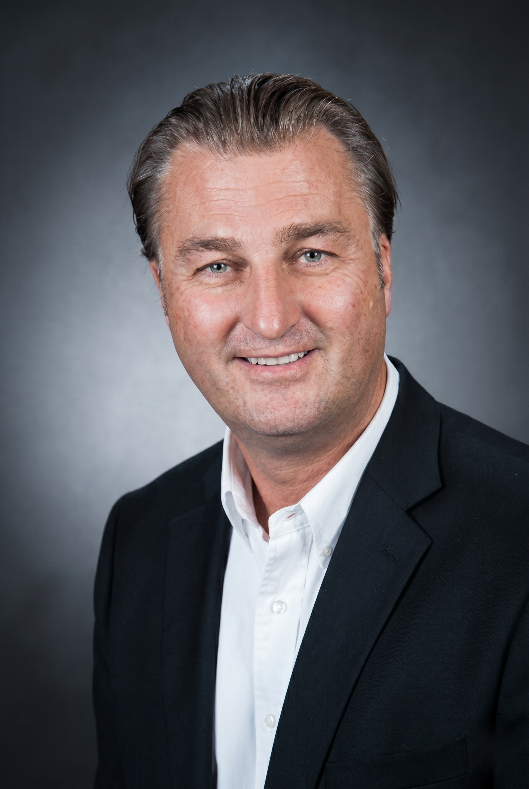 Der Mann trägt einen schwarzen Anzug und ein weißes Hemd und lächelt leicht, vor einem grauen Hintergrund.