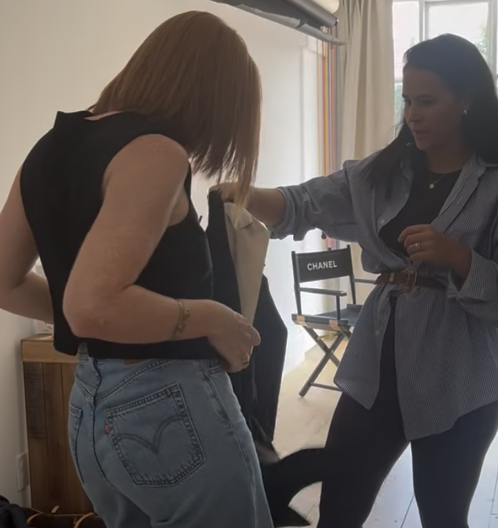Two women in casual clothing interacting with a clothing item in a room with natural light, a Chanel chair, and a small wooden table.