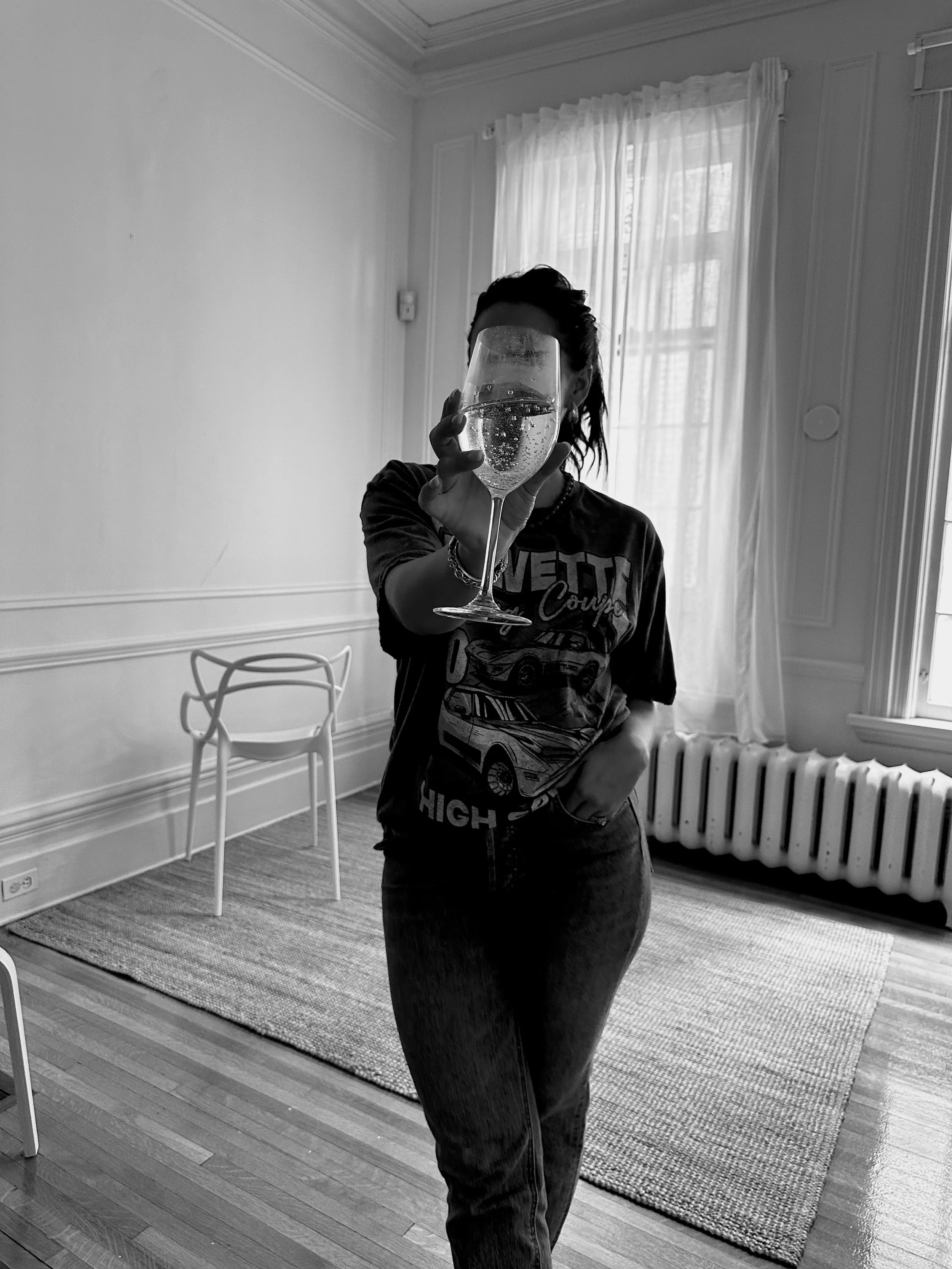 Person holding a tall glass of sparkling water or champagne in front of their face, standing in a well-lit room with curtains and wooden flooring.