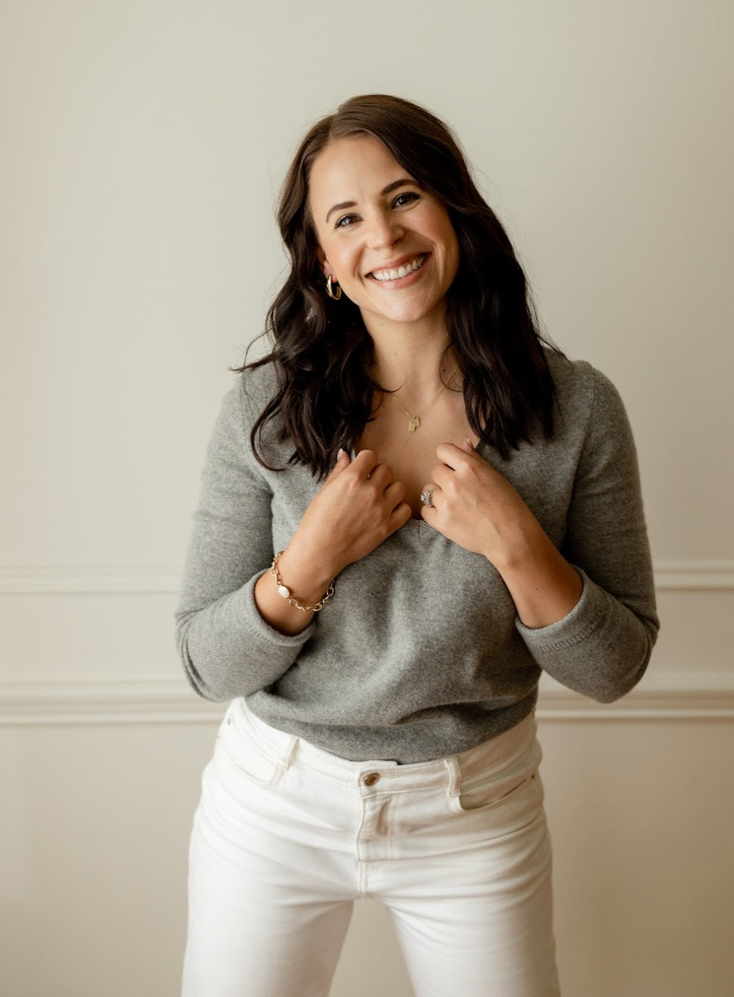 A woman with shoulder-length dark wavy hair smiling, wearing a gray sweater and white pants, accessorized with jewelry, standing in front of a plain off-white wall.