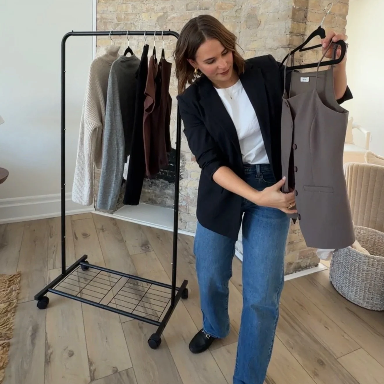 Woman looking at a gray sleeveless dress on a hanger, standing in a room with a clothing rack holding various garments.