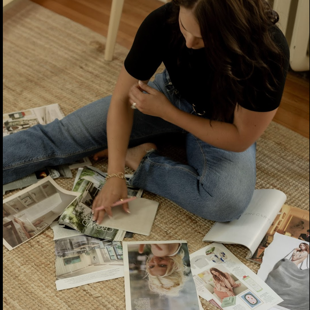 A woman sitting on a woven rug, looking at and sorting through various magazines and catalogs with interior design and fashion images.