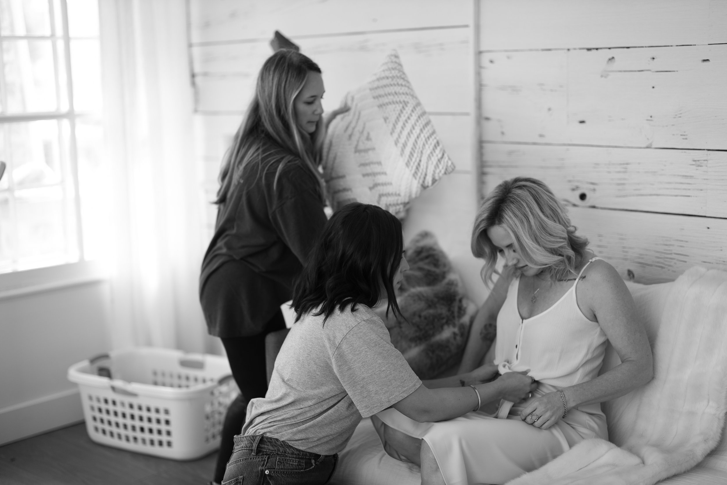 A woman in a wedding dress is seated, surrounded by four people, with one standing and three sitting close to her, possibly during a wedding preparation or fitting.