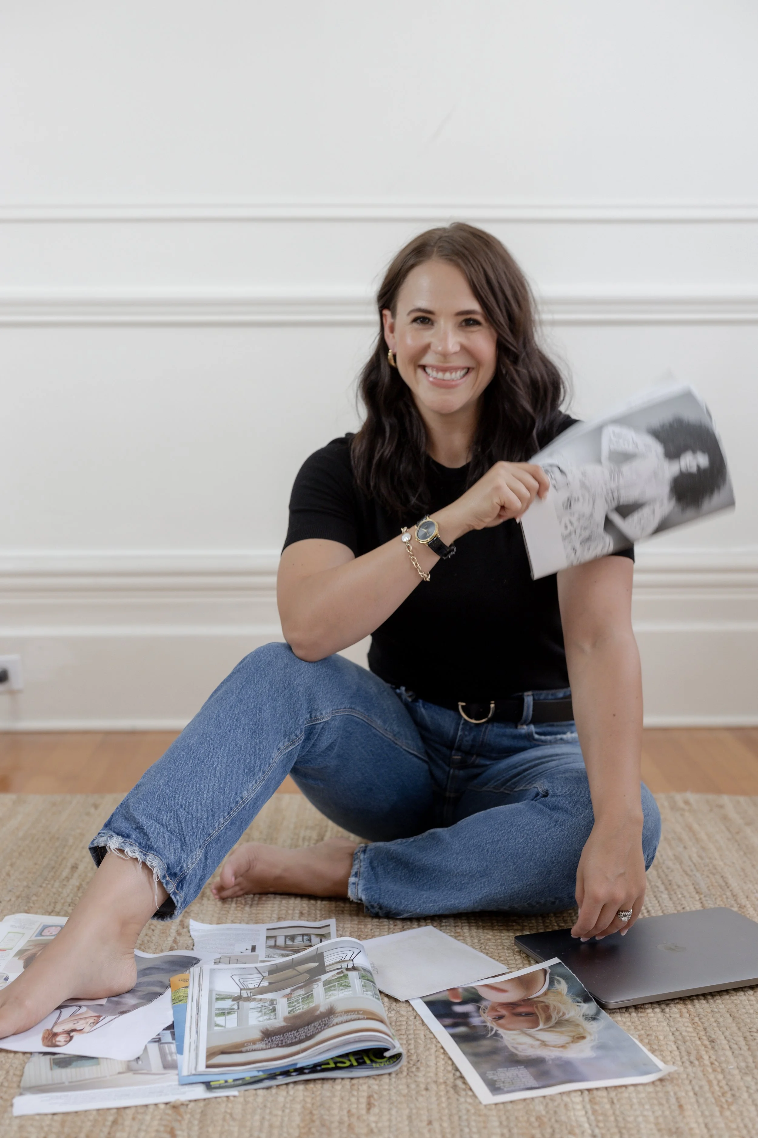 A woman with wavy brown hair smiling while sitting on the floor with magazines, papers, a laptop, and a tablet spread around her.