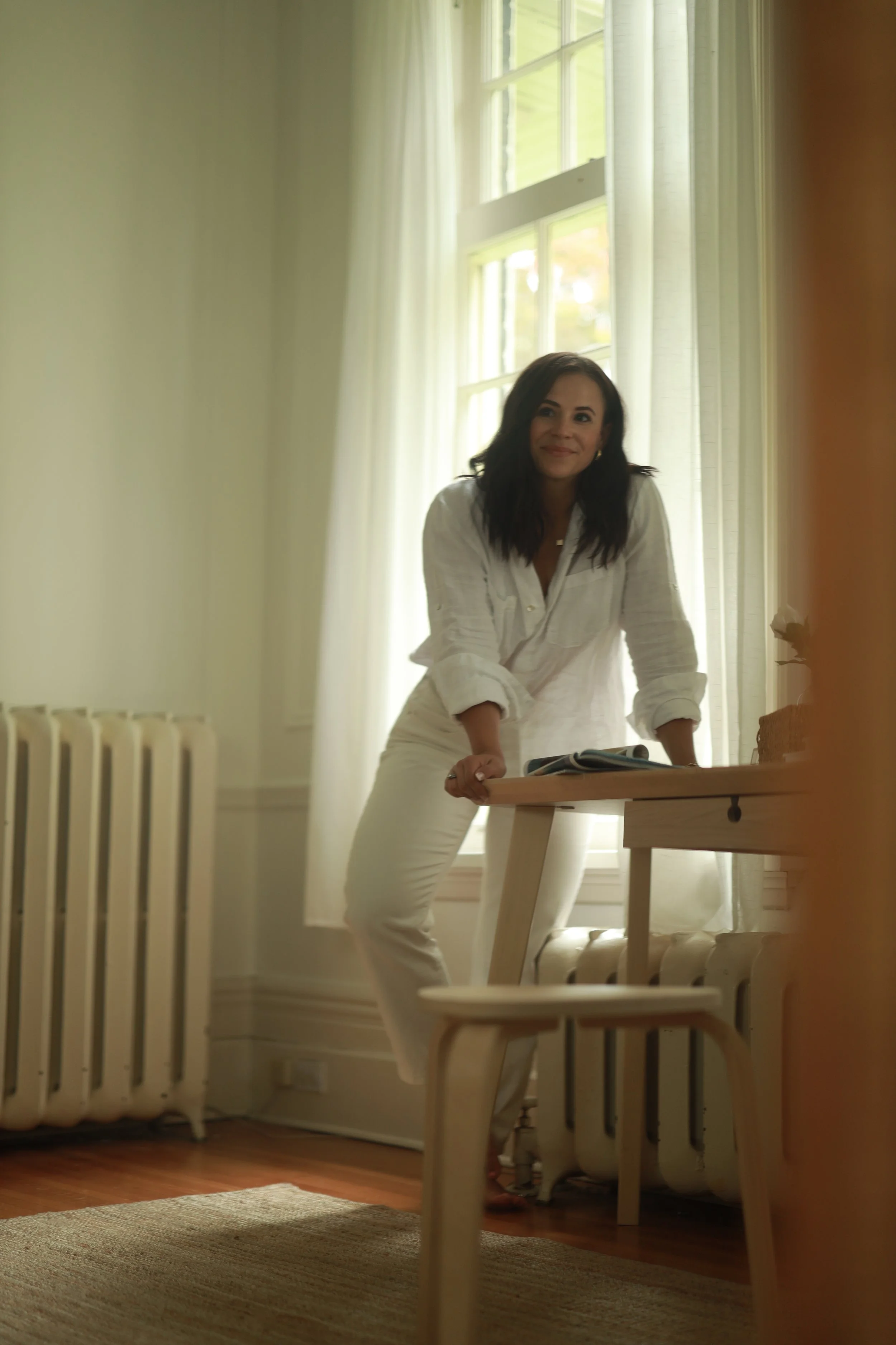 A woman in a white shirt and white pants leaning on a wooden table near a window with curtains, in a well-lit room with hardwood floors and a beige rug.
