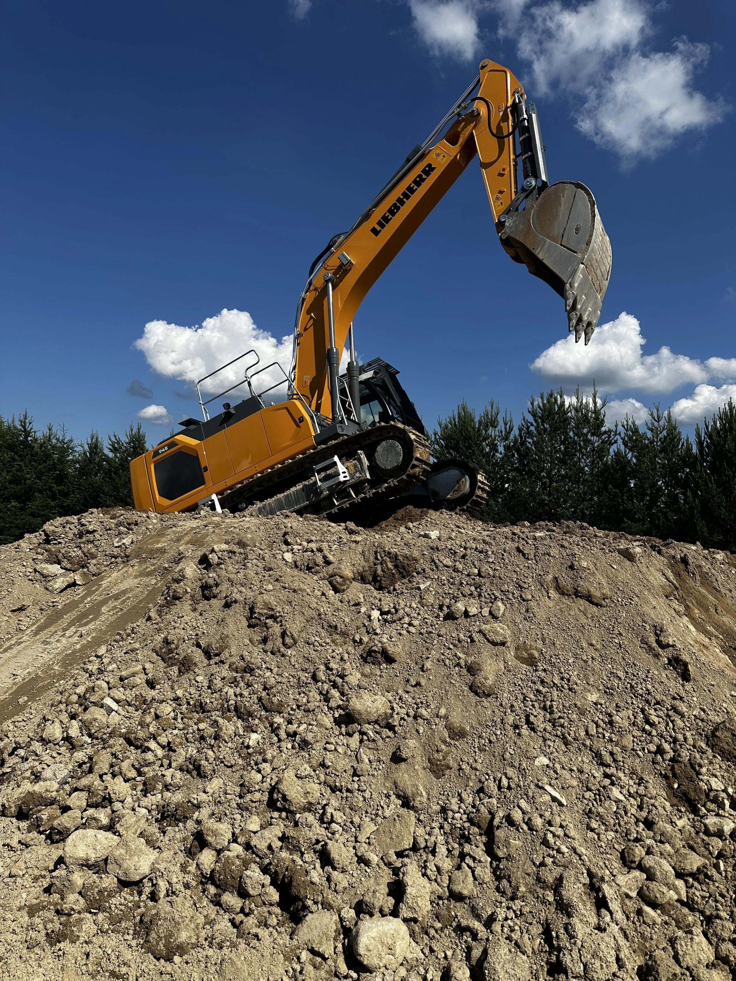 Une excavatrice jaune en action déplaçant de la terre à ciel bleu avec quelques nuages et des arbres en arrière-plan.