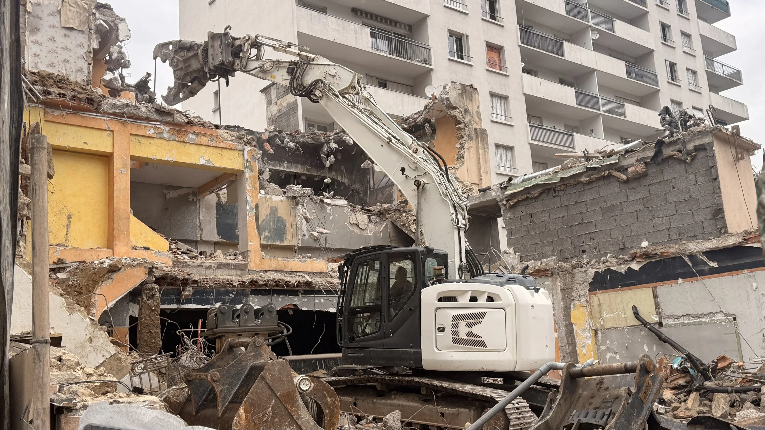 Une excavatrice en train de détruire un bâtiment d'appartements en béton, avec des débris et des murs en ruine autour.