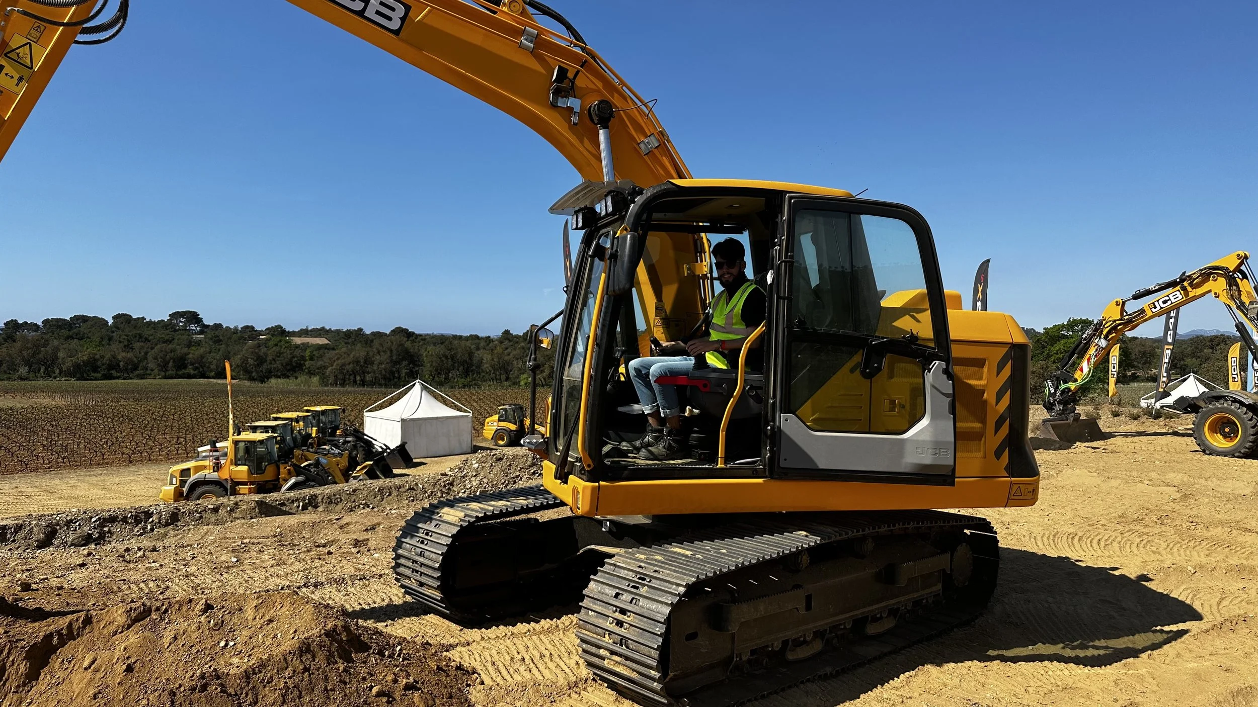Une excavatrice jaune avec un homme dans la cabine portant une veste fluorescente, travaillant sur un terrain de terre. D'autres excavatrices jaunes et une tente blanche sont visibles dans un champ entouré d'arbres, sous un ciel clair.