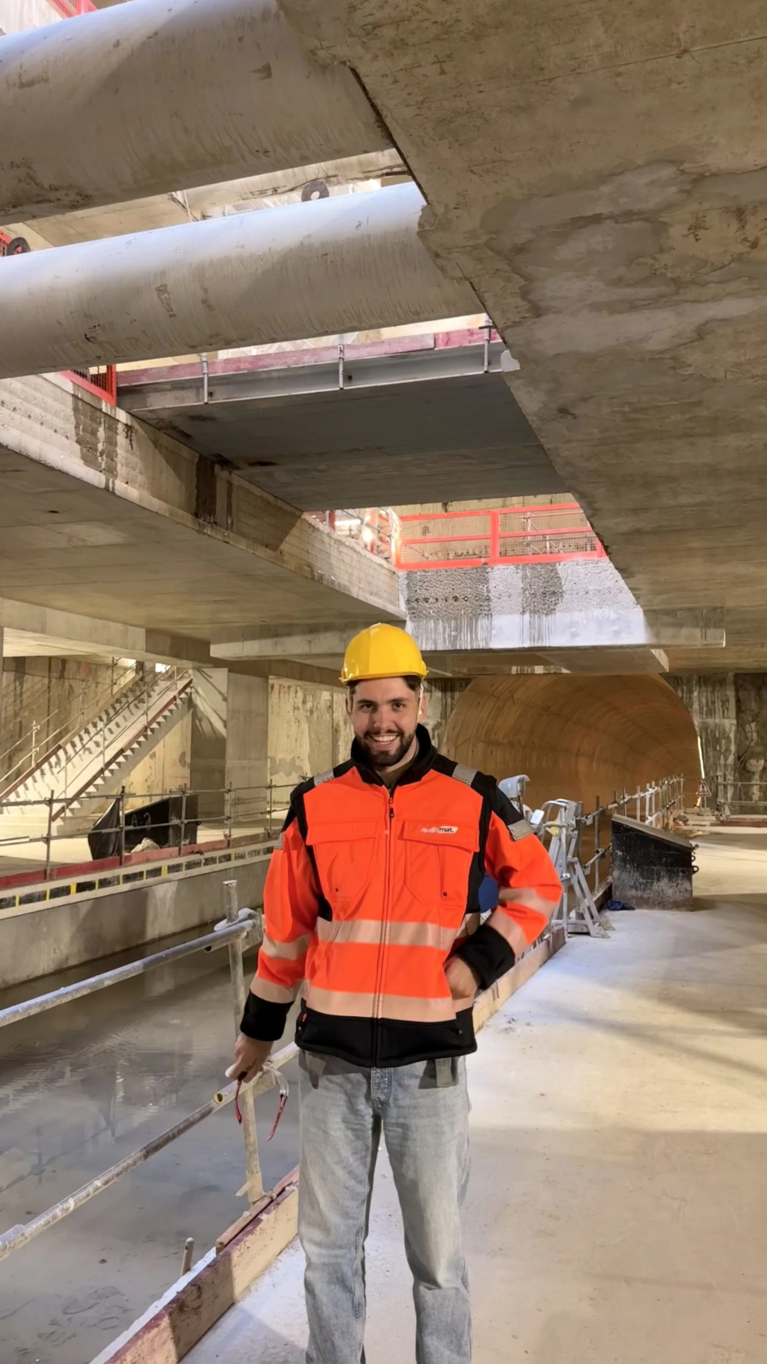 Un homme souriant portant un casque jaune et une veste de chantier orange se tient dans un tunnel en construction à l'intérieur d'une infrastructure.