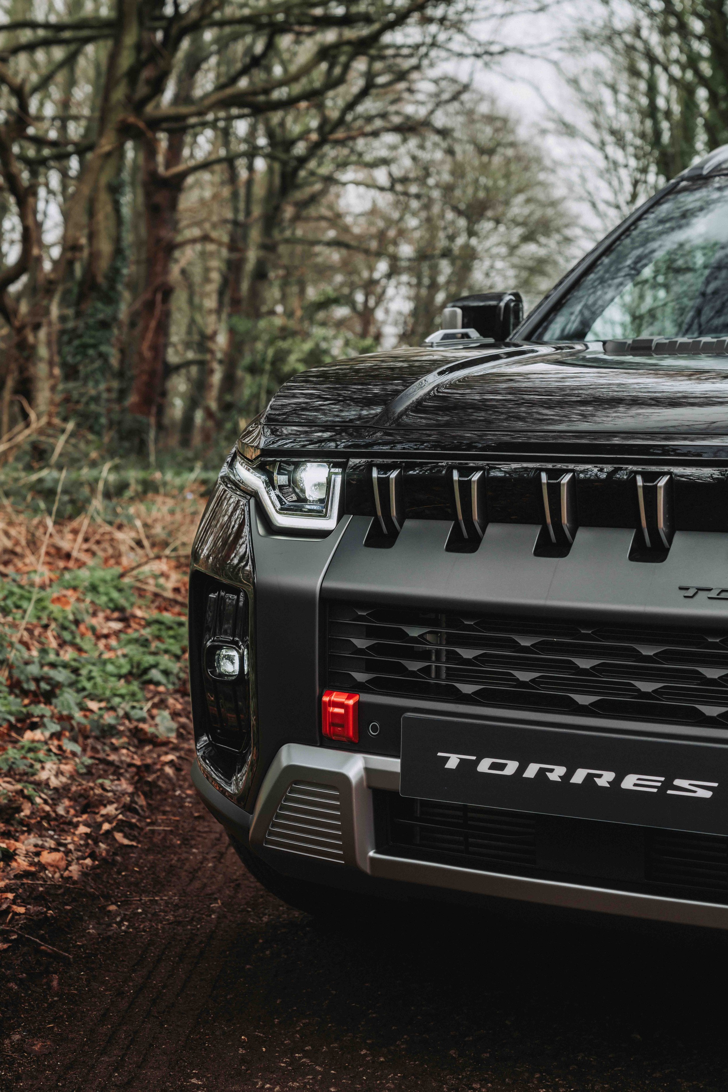 Close-up of the front right side of a black and gray TORRES SUV parked on a forest trail with trees and leaves in the background.