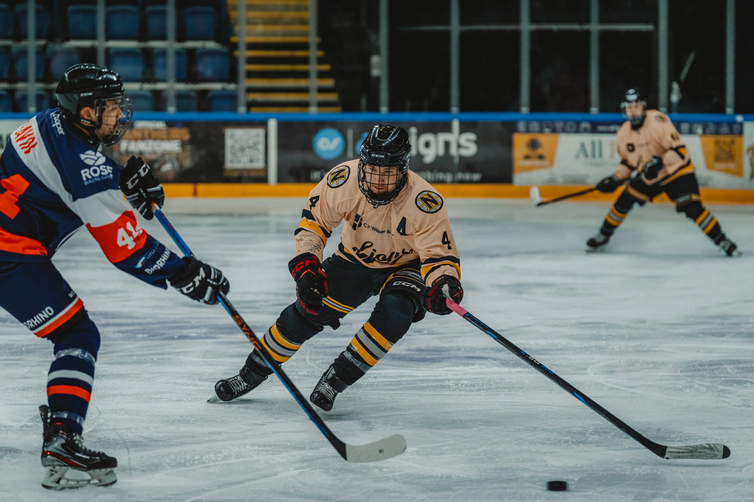 Ice hockey game with players in action on the rink, including one in a yellow jersey controlling the puck and others nearby.