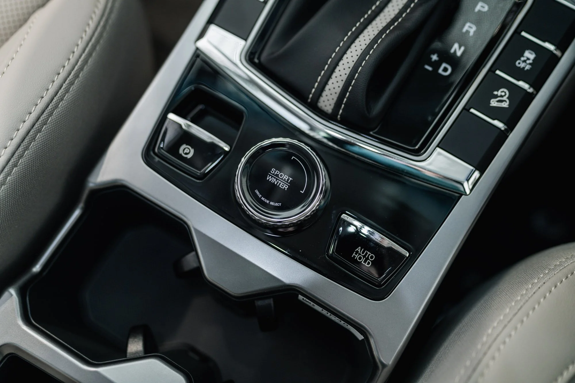 Close-up of a car's center console featuring a gear shifter, a drive mode selector dial labeled 'Sport Winter' with options for drive mode select, and buttons for parking brake, auto hold, and other functions.