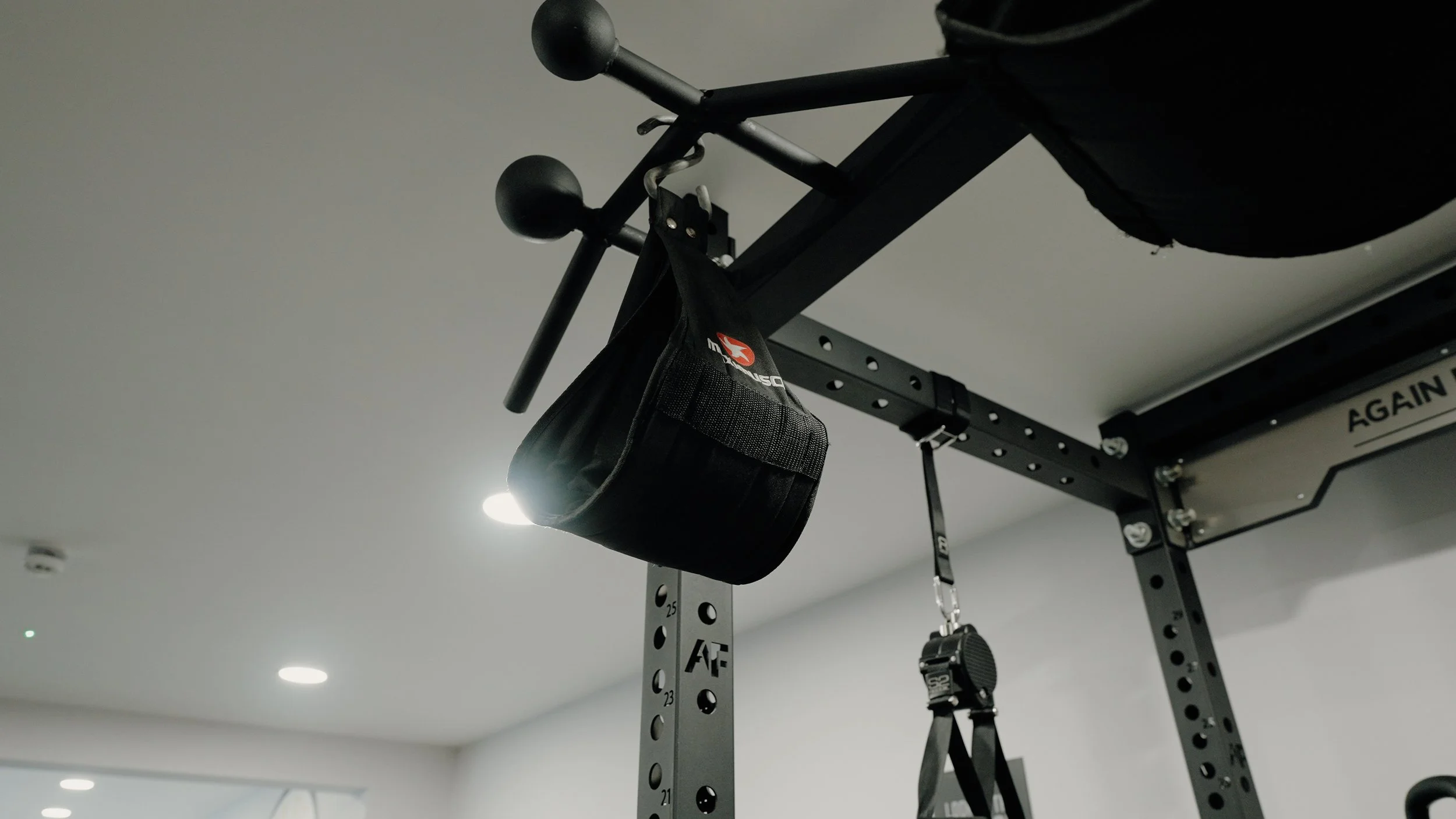 Black gym equipment with a small black pouch hanging from a bar and a black strap with a round adjustment knob hanging underneath, inside a gym with ceiling lights.